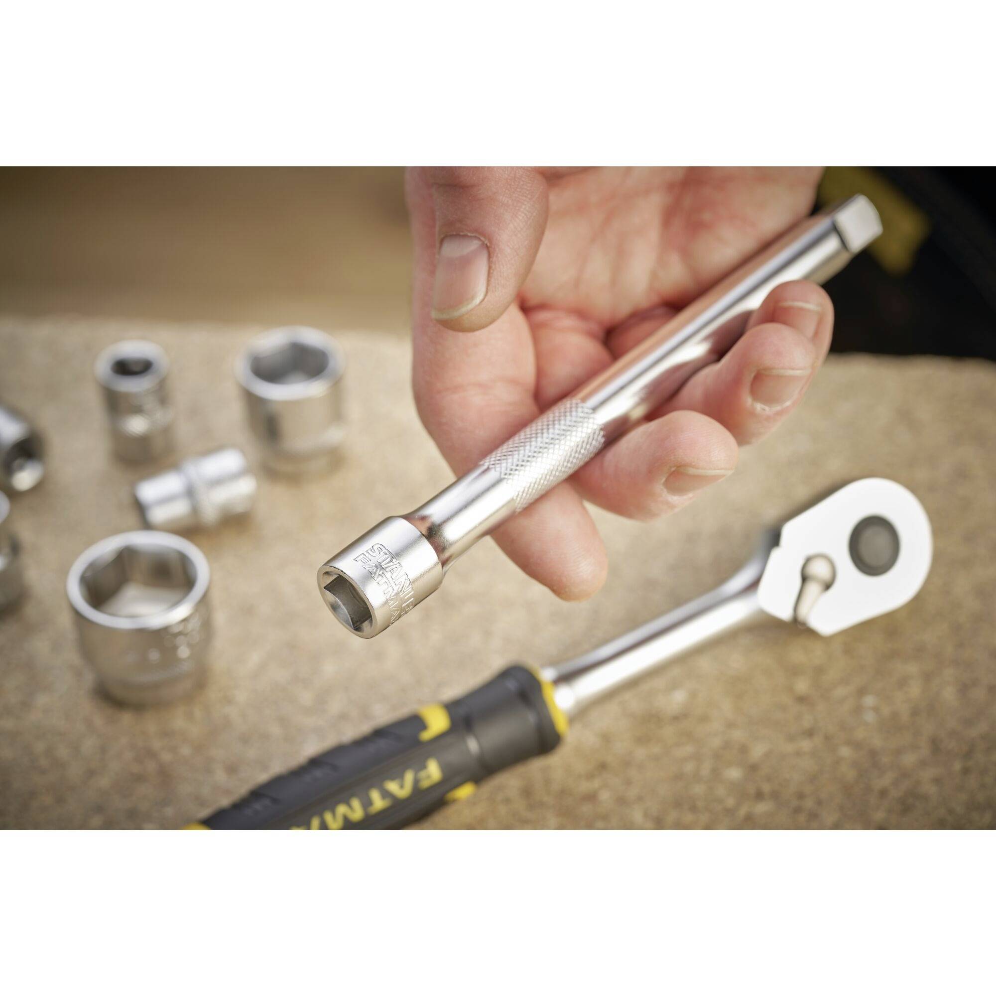 A hand is holding a silver socket extension bar. Beside it are a spanner and various socket attachments.