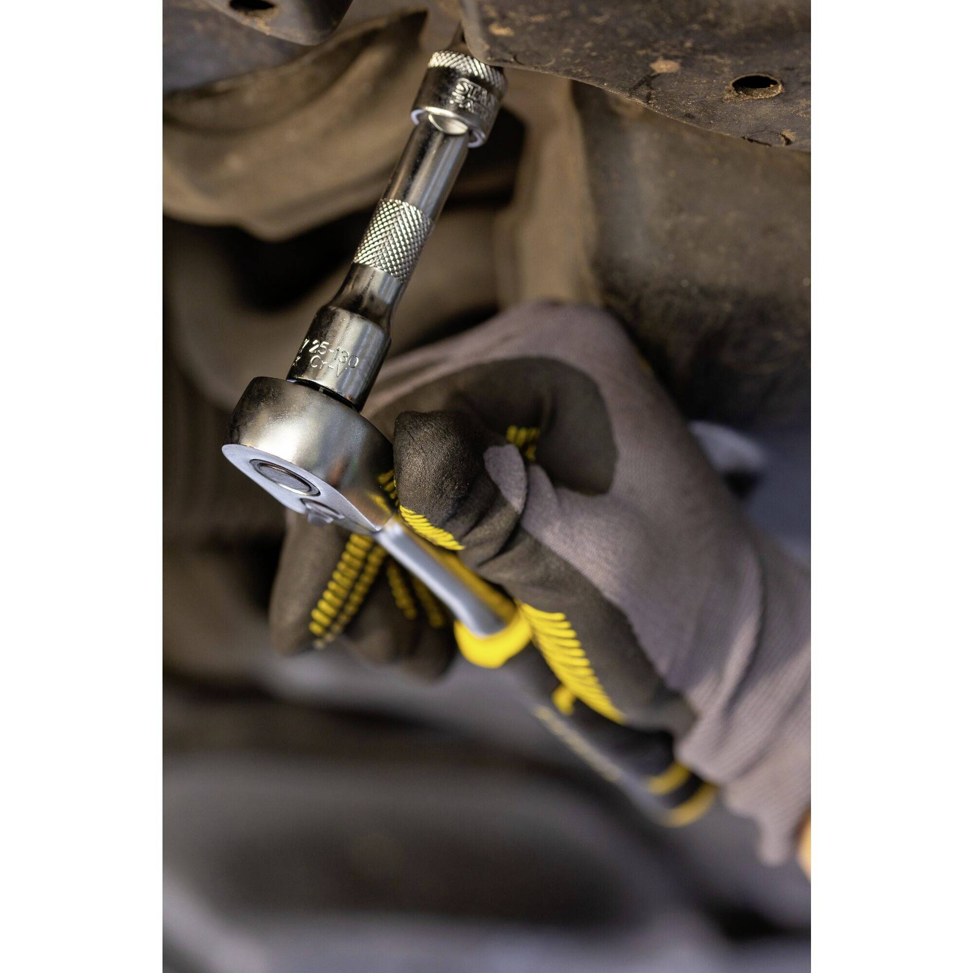 A hand wearing a grey glove holds a ratchet spanner and tightens a screw while working underneath a vehicle.