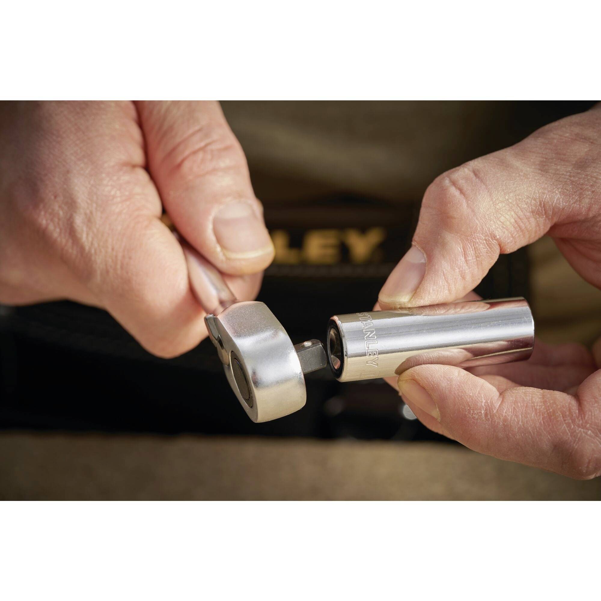 Two hands are operating a silver tool. One hand is holding the handle while the other turns the spanner head. Equipment is visible in the background.