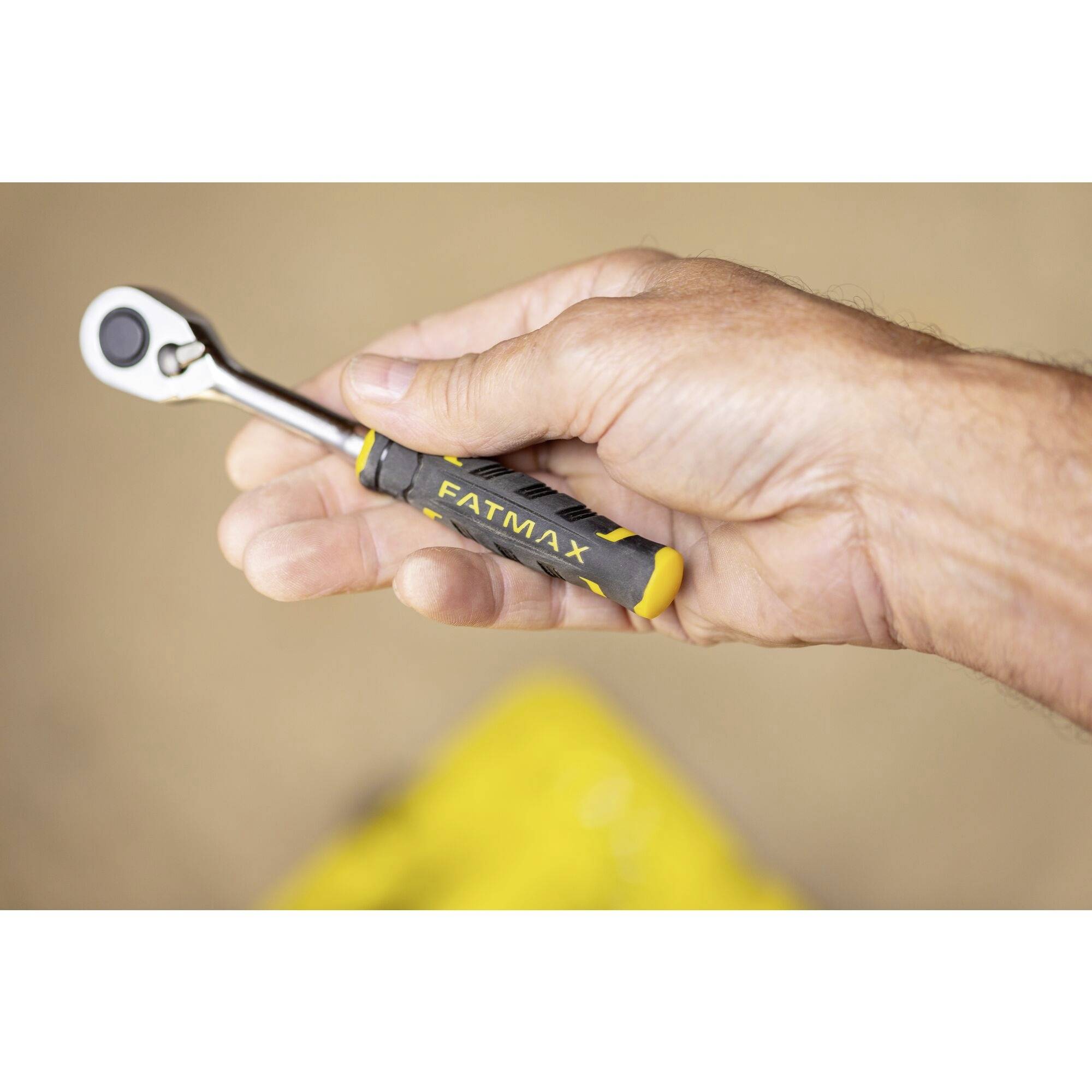 A hand is holding a ratchet spanner with a yellow and black handle, with 'FATMAX' printed on it. The background is out of focus.