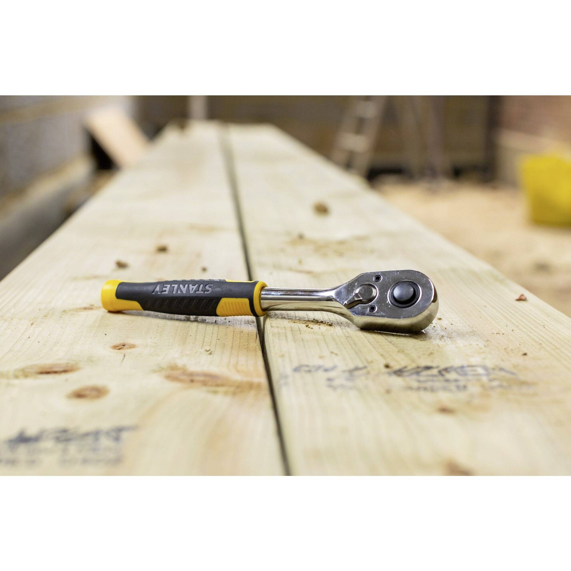 A ratchet with a yellow and black handle lies on a wooden beam in a workshop. The background is out of focus.