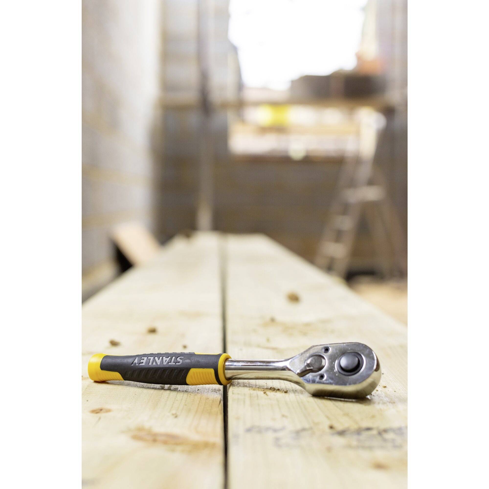 A yellow-handled ratchet lies on a wooden beam at a construction site. A ladder is visible in the background.