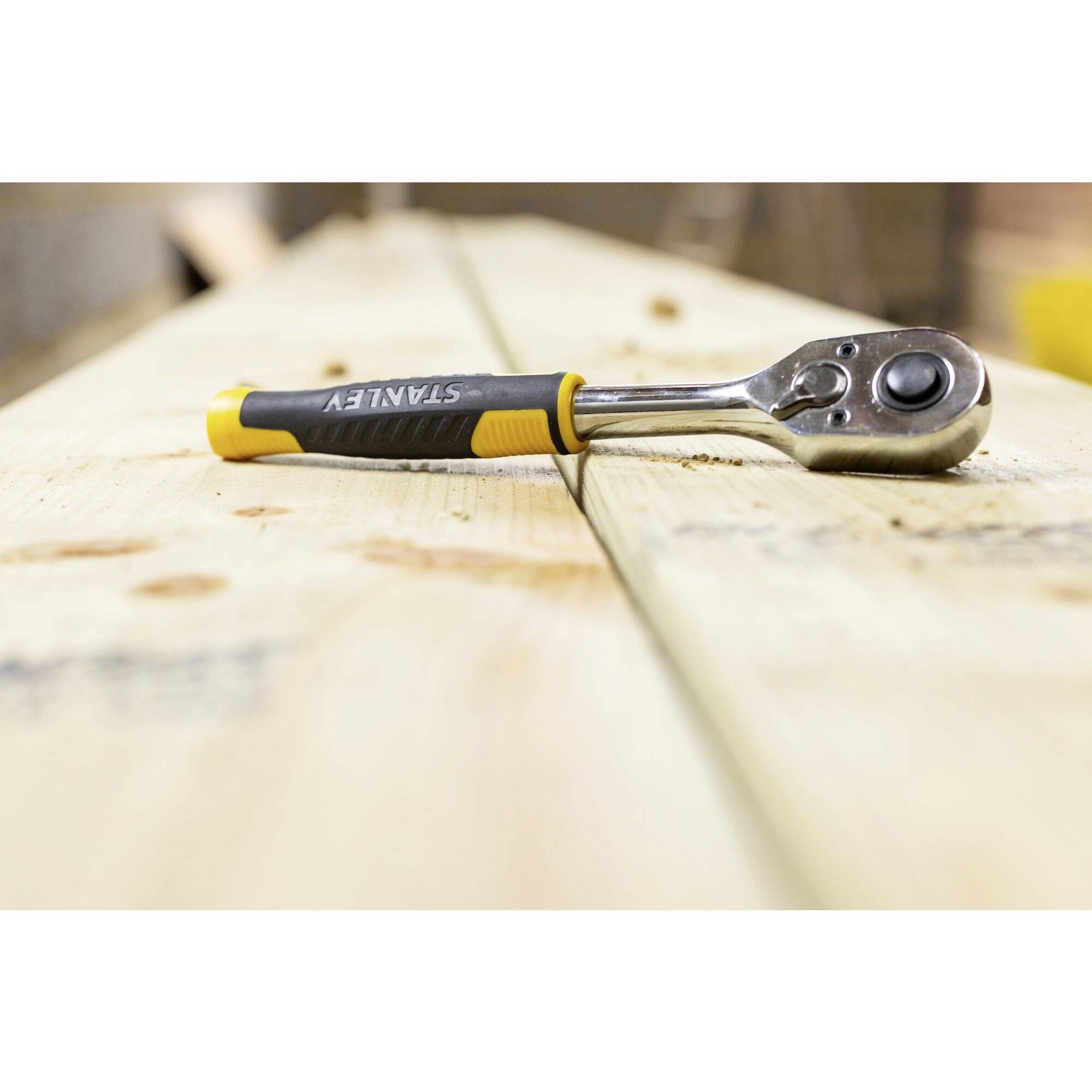 A ratchet spanner lies on a wooden beam in a workshop. The tool has a black and yellow handle.