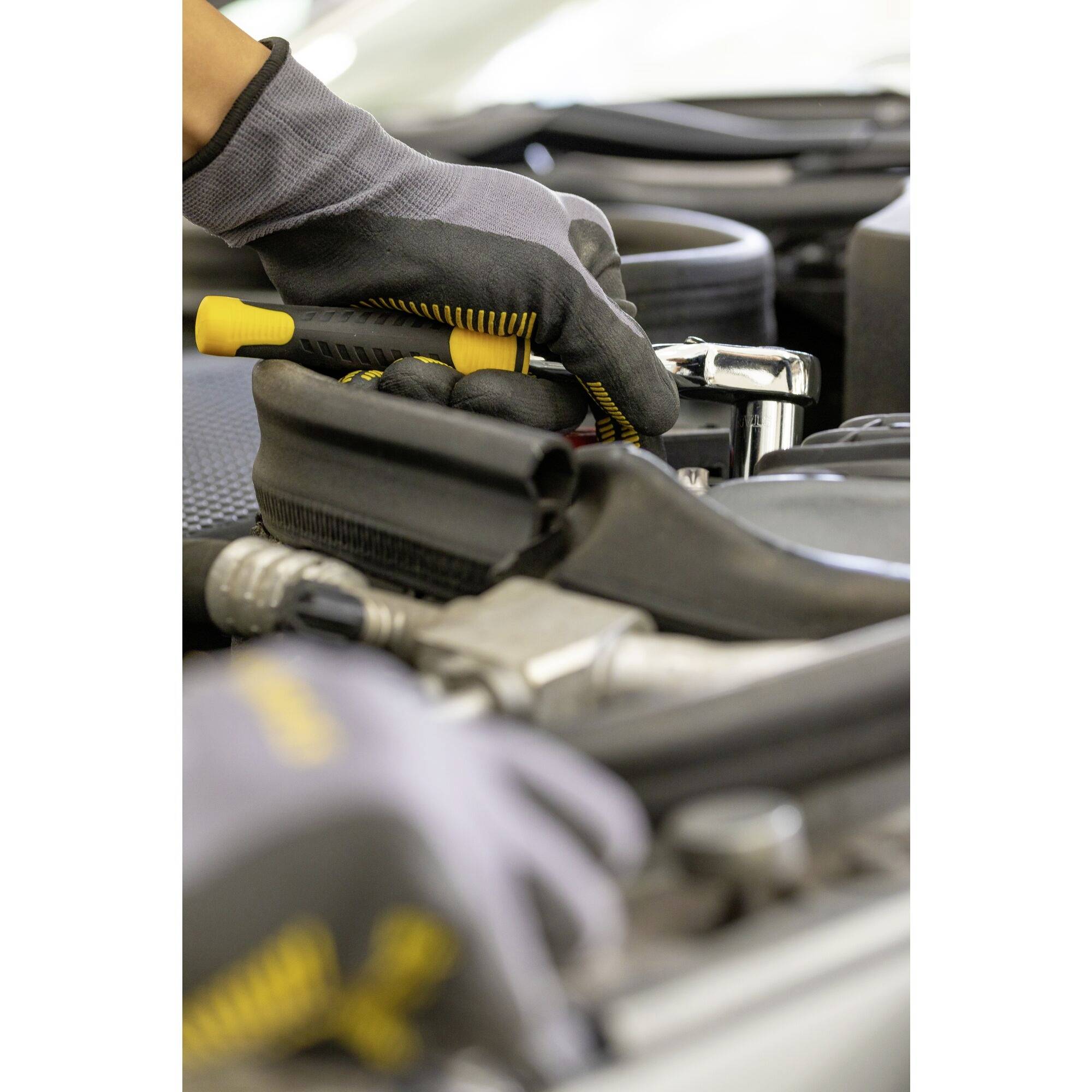A mechanic wearing grey gloves is tightening a bolt with a spanner in the engine compartment of a car.