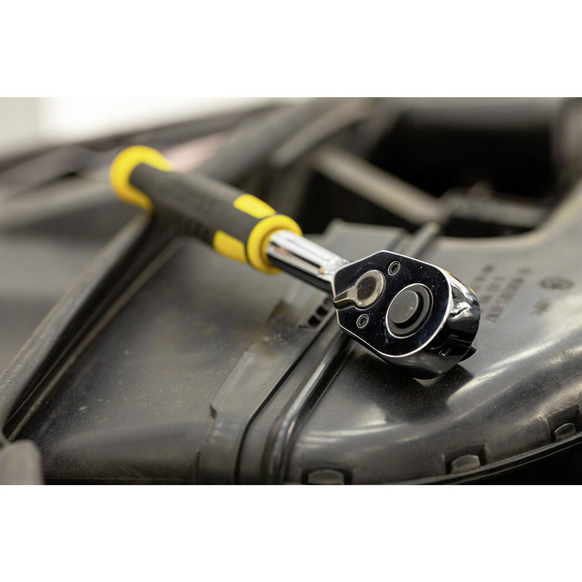 A chrome-plated ratchet with a yellow handle lies on a black engineering component. The ratchet is positioned slightly at an angle in the foreground.