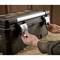 A person opens a large black toolbox with silver latches. The box is sitting on a table covered with sawdust.