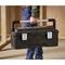 A person is holding a large black toolbox with a metal handle and two latches. The background shows wooden frames.