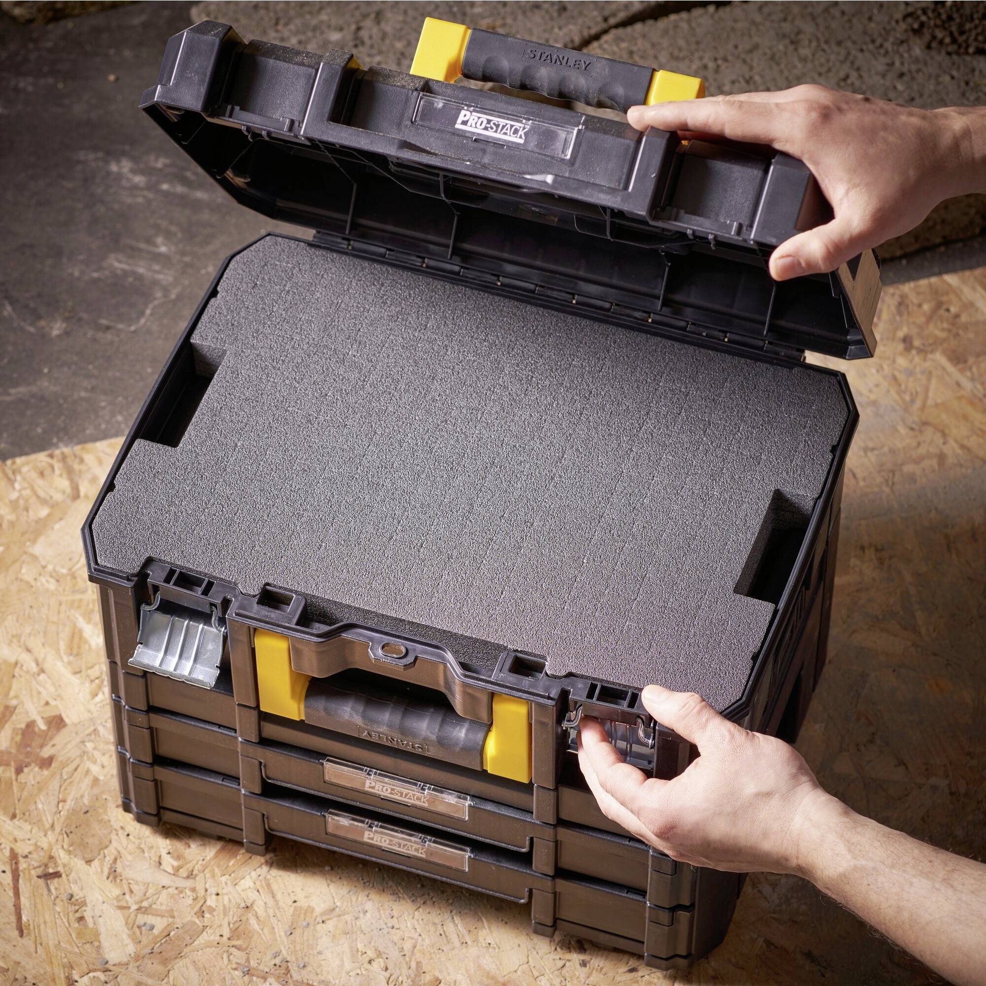 A person opens a black tool case with yellow catches on a wooden floor. The case is empty and made of plastic.
