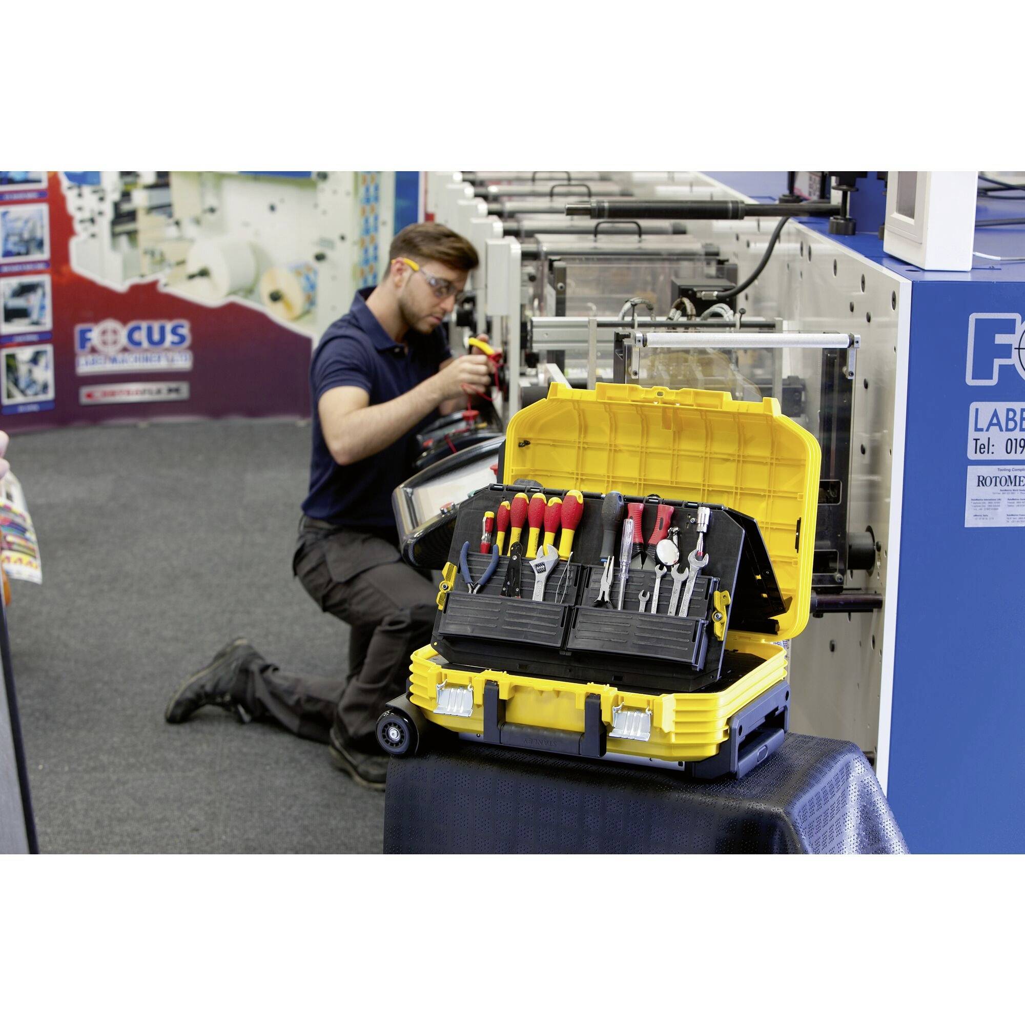 A man is repairing a machine. In the foreground, an open toolbox with various tools. The background shows an industrial setting.