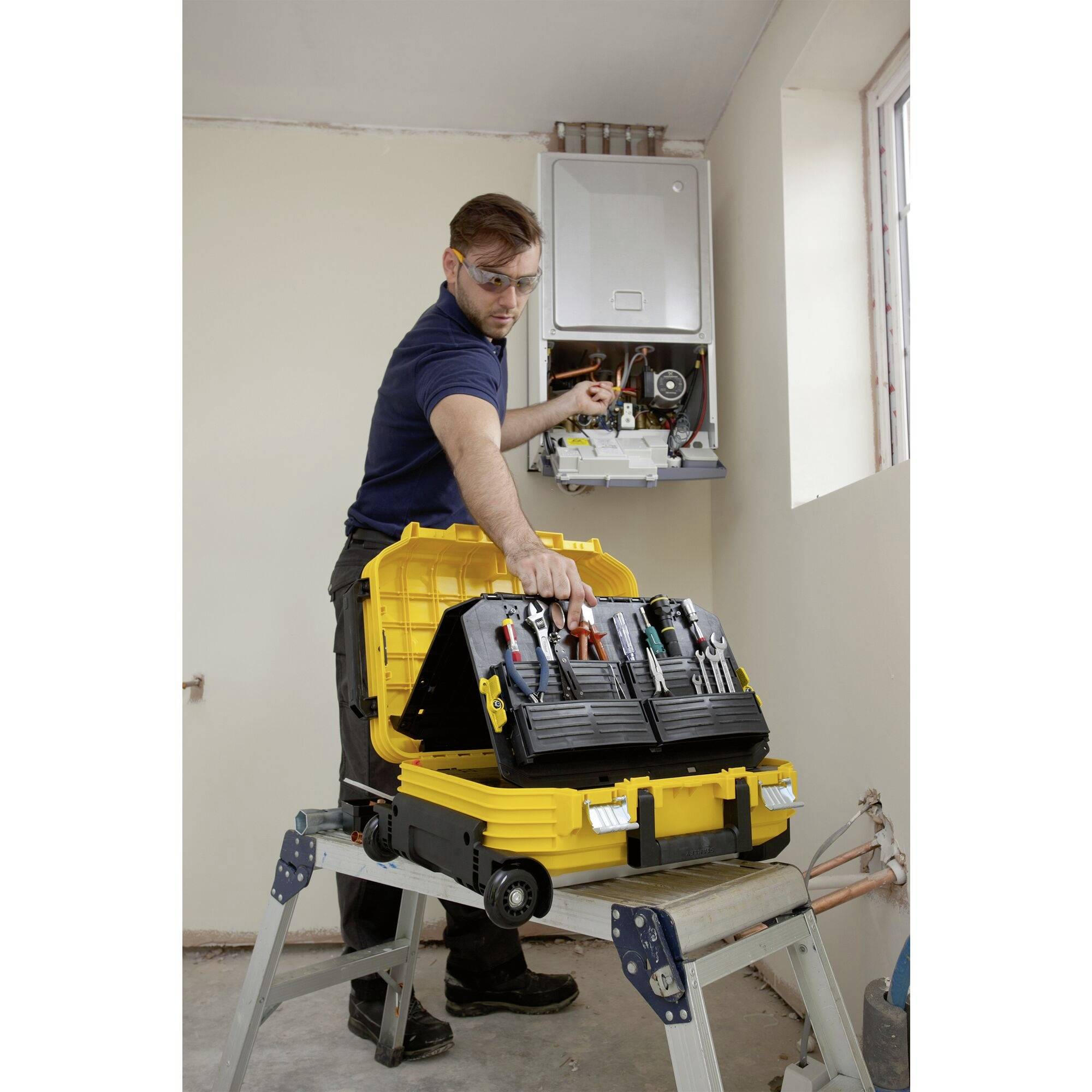 A tradesman is repairing a gas boiler in a room with white-painted walls. A yellow toolbox is sitting on a trestle.