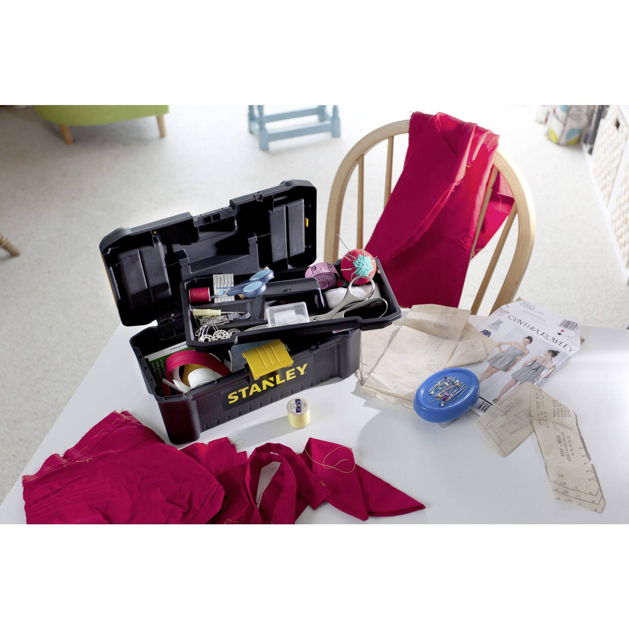 Sewing supplies on a table: An opened toolbox with scissors, tape measure, and threads. Red fabrics, pattern cutting paper, and sewing accessories beside it.