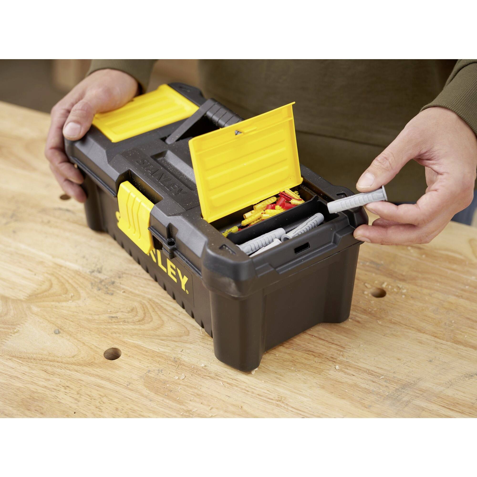 A person opens a black toolbox with a yellow lid and removes tools. The box is sitting on a wooden table.
