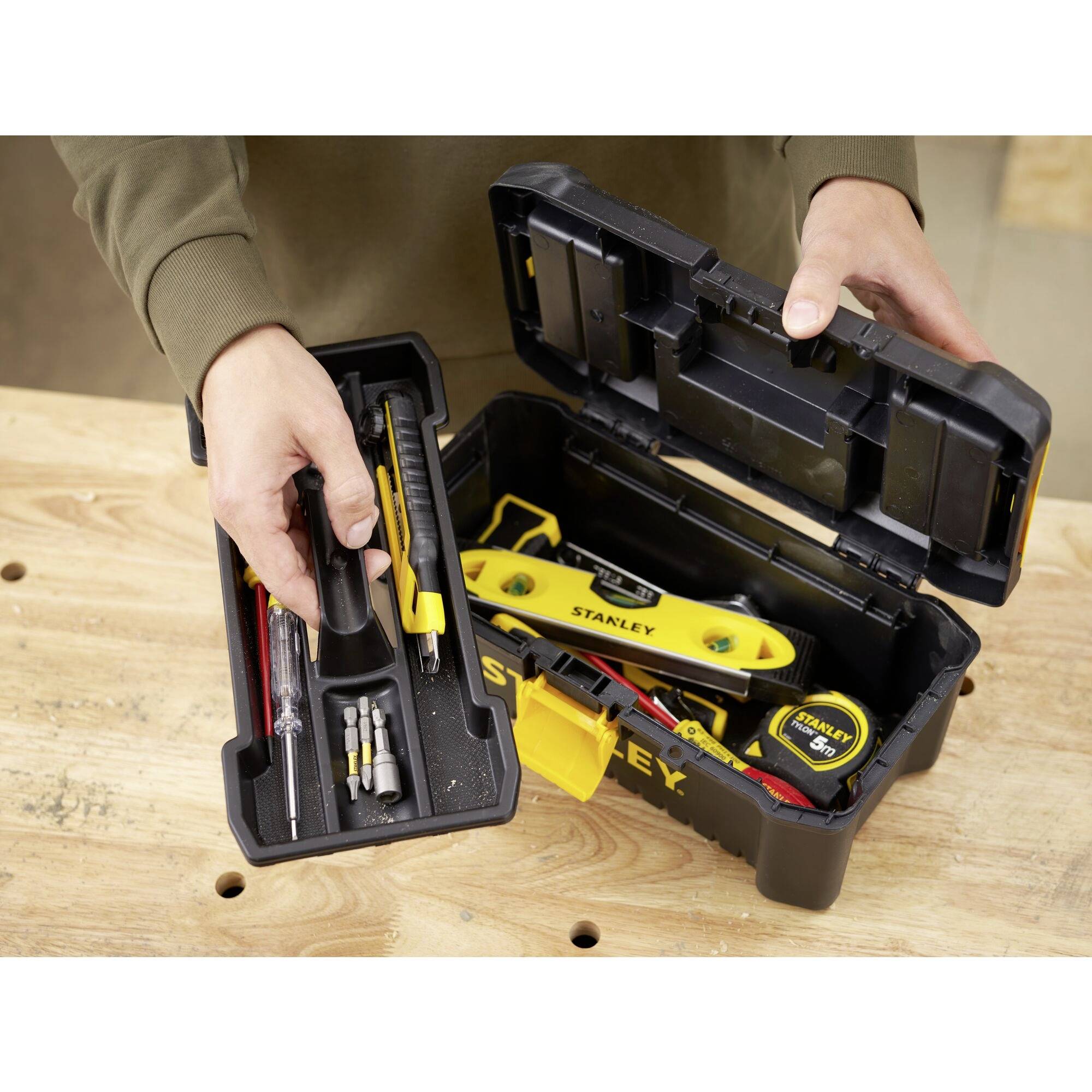 A person opens a toolbox containing screwdrivers, a tape measure, and bits. Everything is lying on a wooden surface.