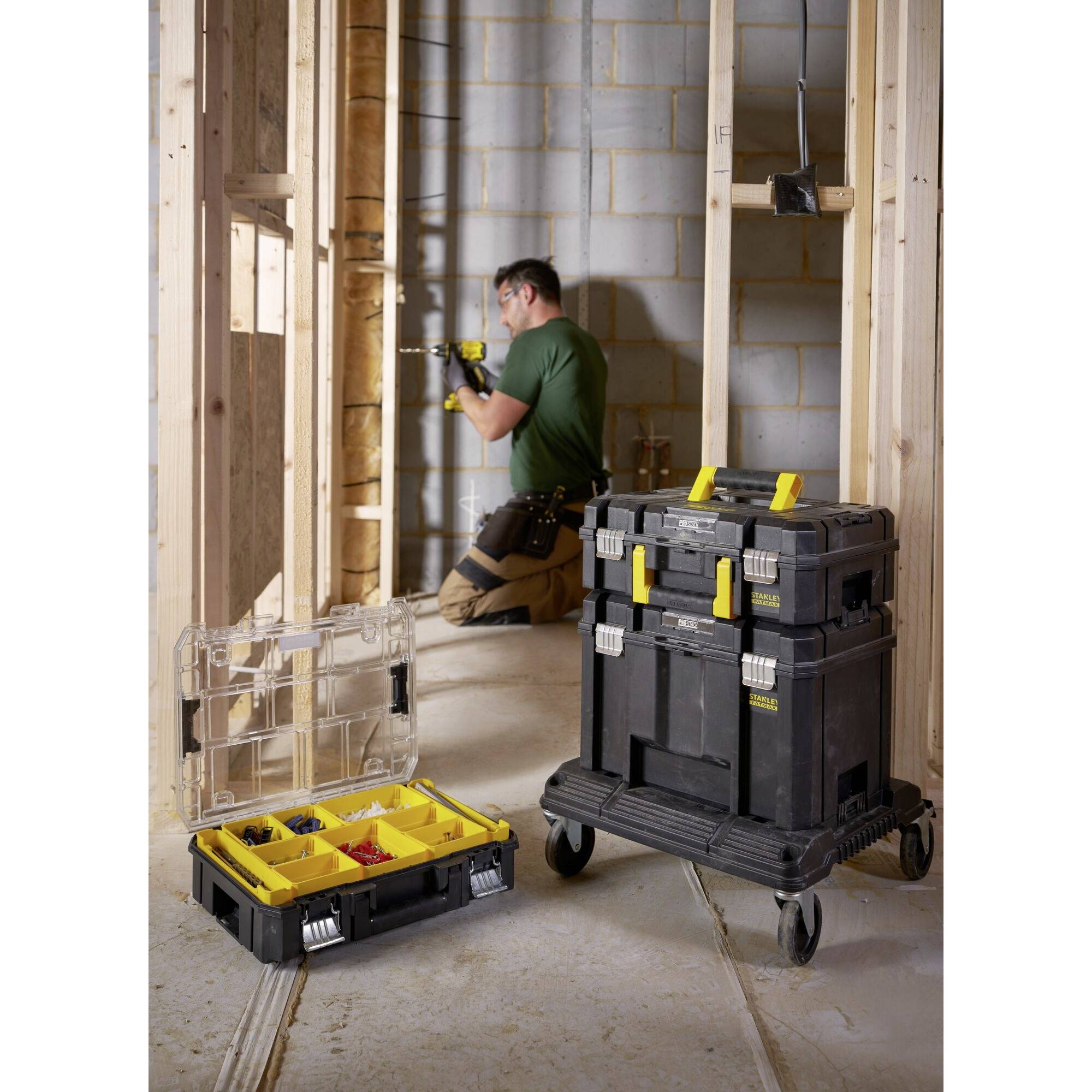 A tradesman is working with tools in an unfinished building. Next to him stands an open toolbox with yellow compartments.