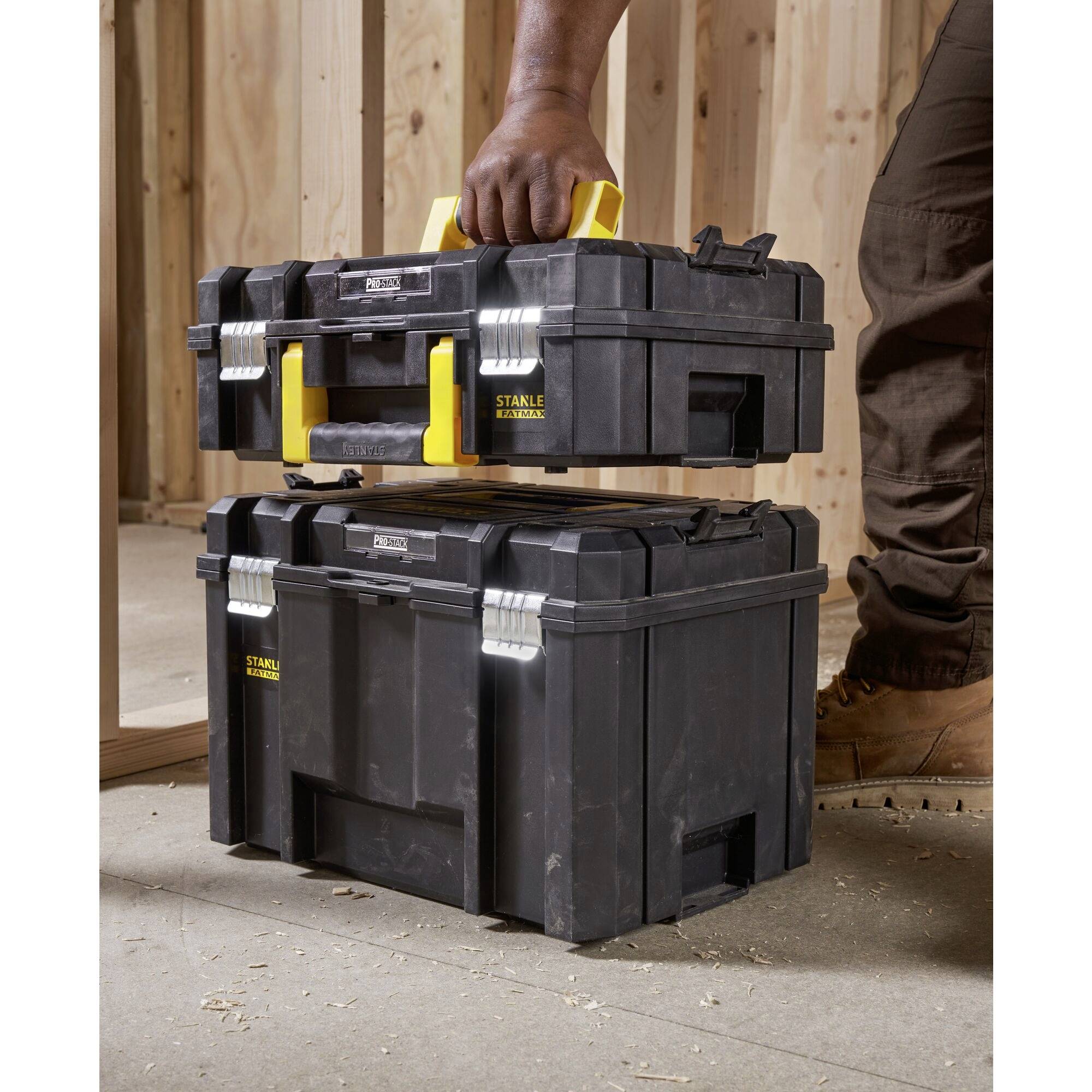 A person lifts a black toolbox with a yellow handle from a stack. The toolbox is situated in a wooden frame structure.