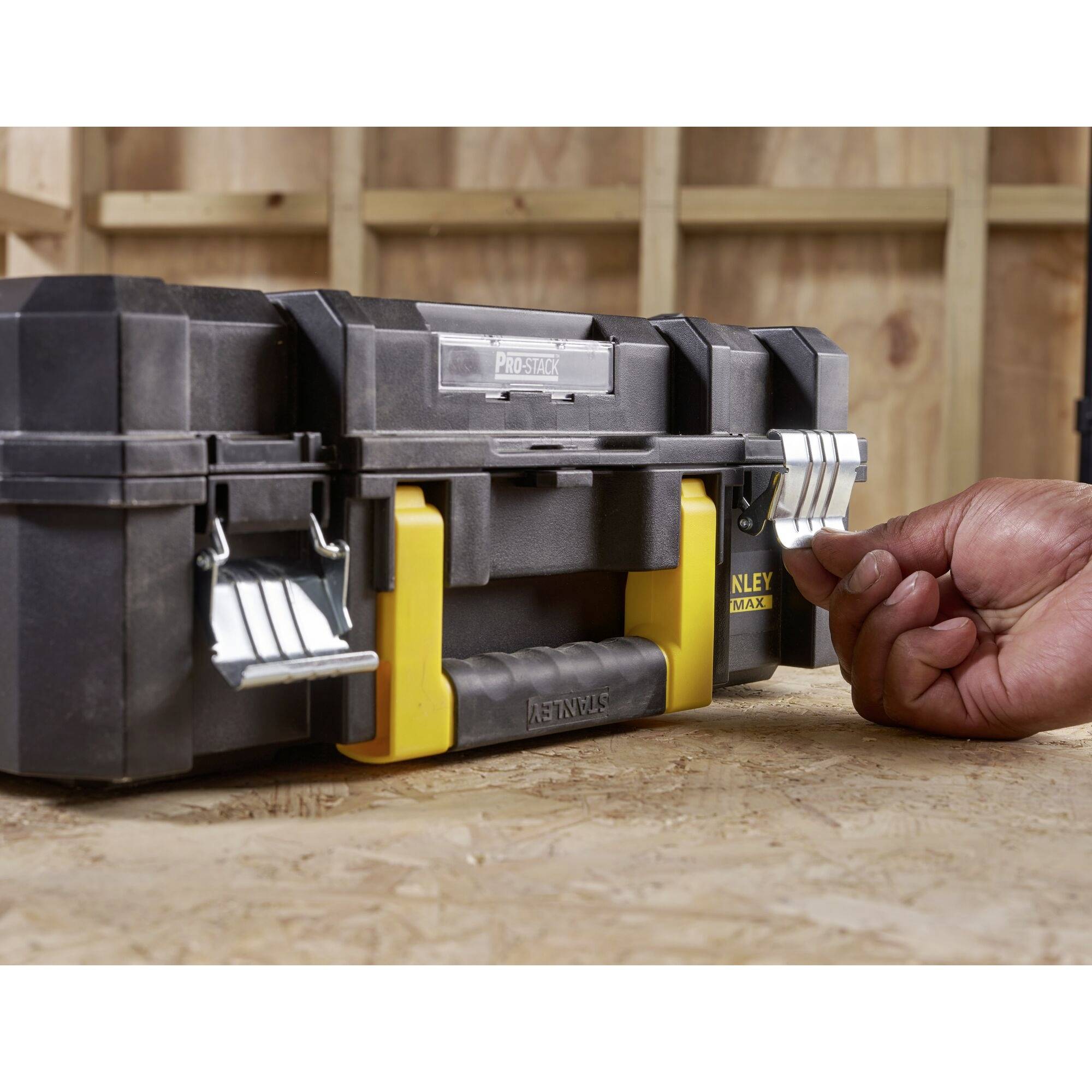 Black toolbox with yellow handle. One hand opens the latch. Background: wooden wall.