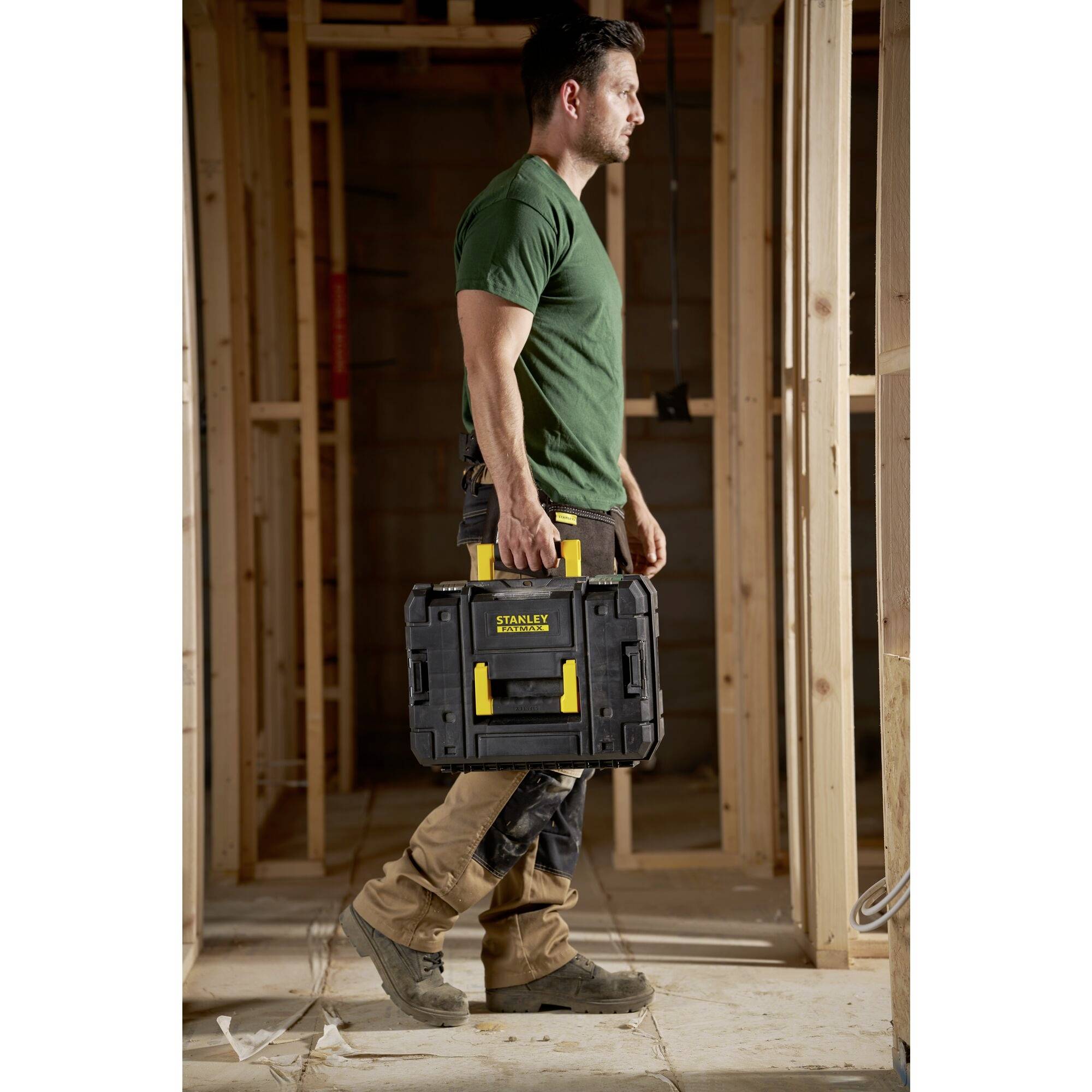 A man is carrying a black toolbox in an unfinished wooden beam framework. He is wearing a green t-shirt and beige trousers.
