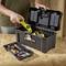 A person places a green and yellow tool into an open black toolbox with a 'Stanley' label on a wooden table.
