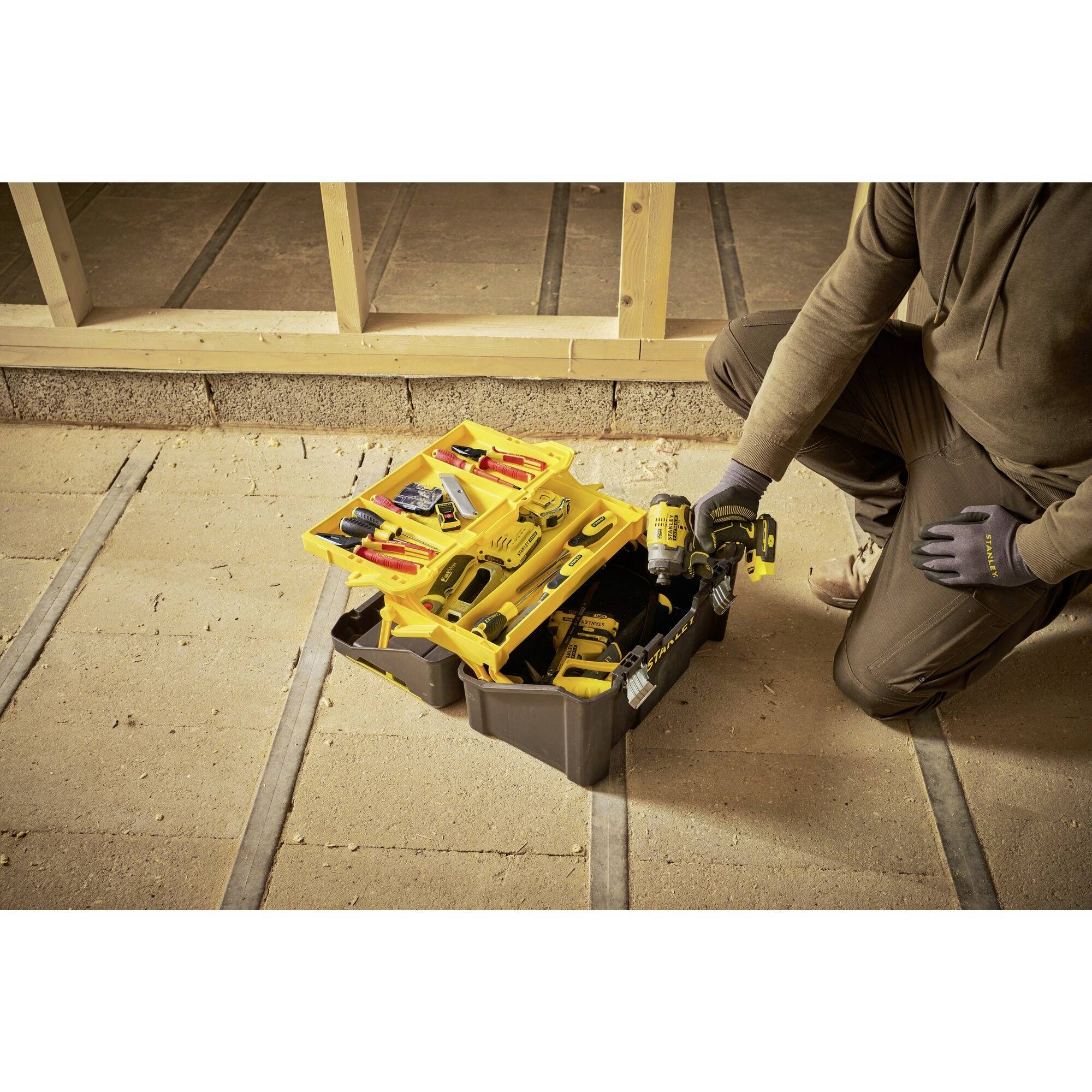A person in work attire is kneeling on a wooden floor and reaching for an electric tool from an open toolbox.