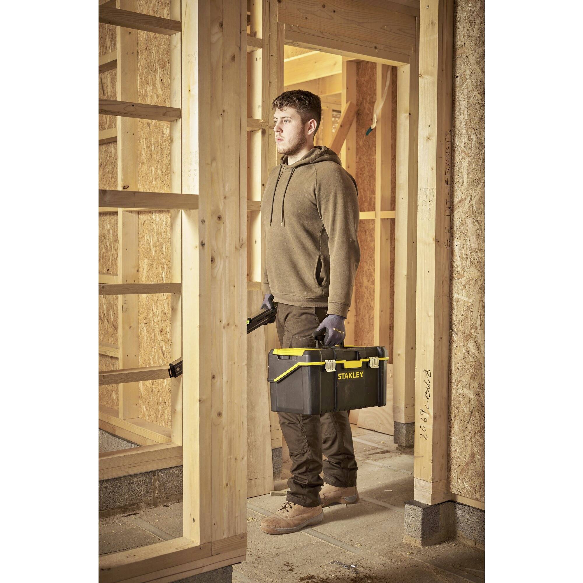 A man in workwear is carrying a toolbox through an unfinished timber-frame construction, as if preparing for building work.