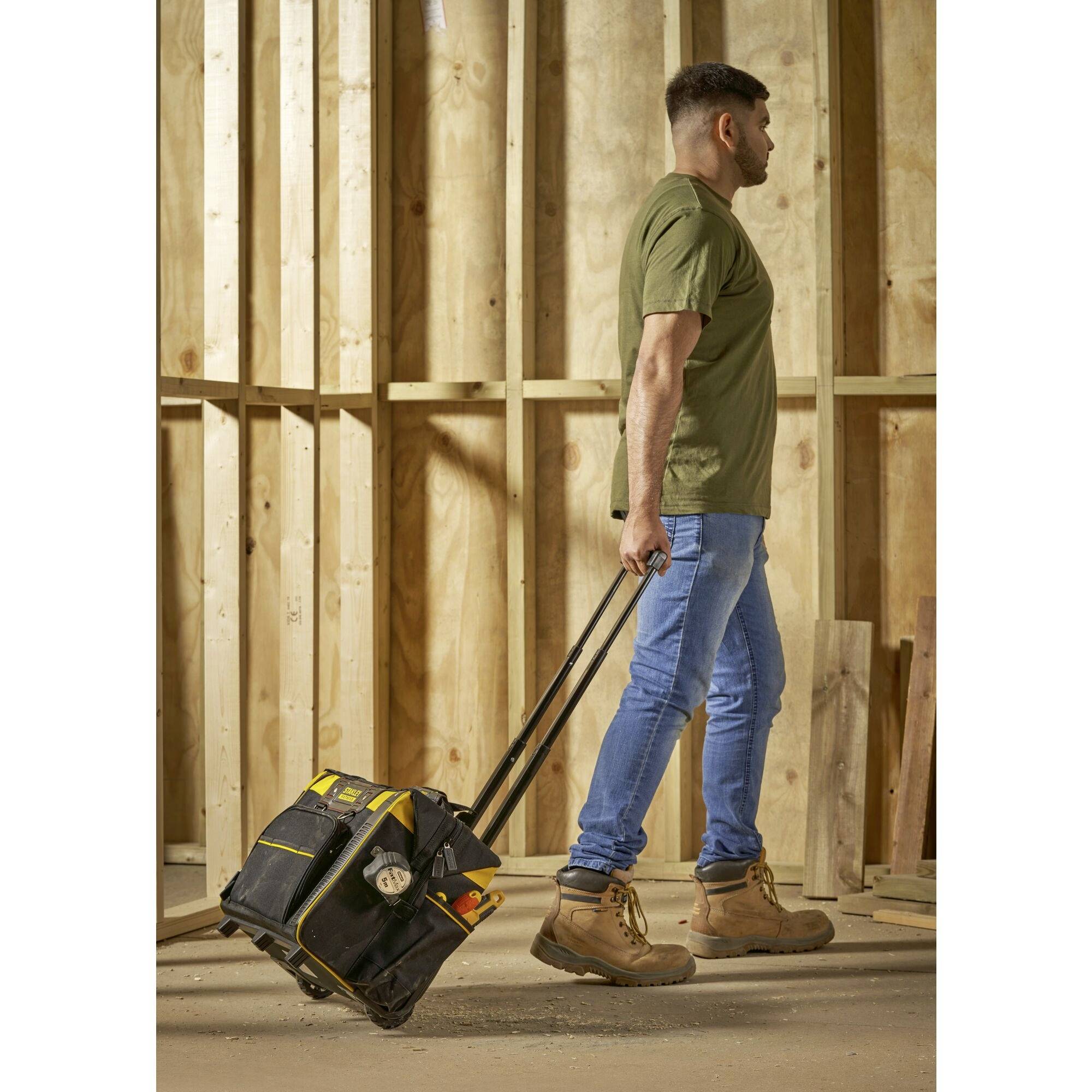A person pulls a tool bag on wheels through a construction site. In the background, wooden wall structures are visible.