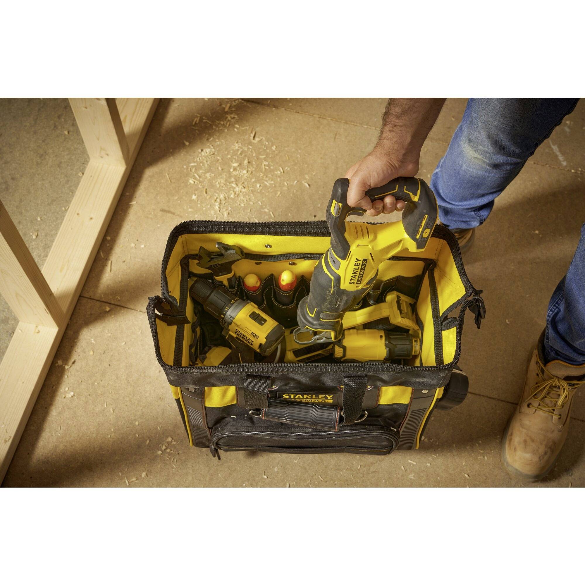 A person is reaching for an electrical tool in a yellow tool bag, which contains other tools. The scene is taking place on a construction site.