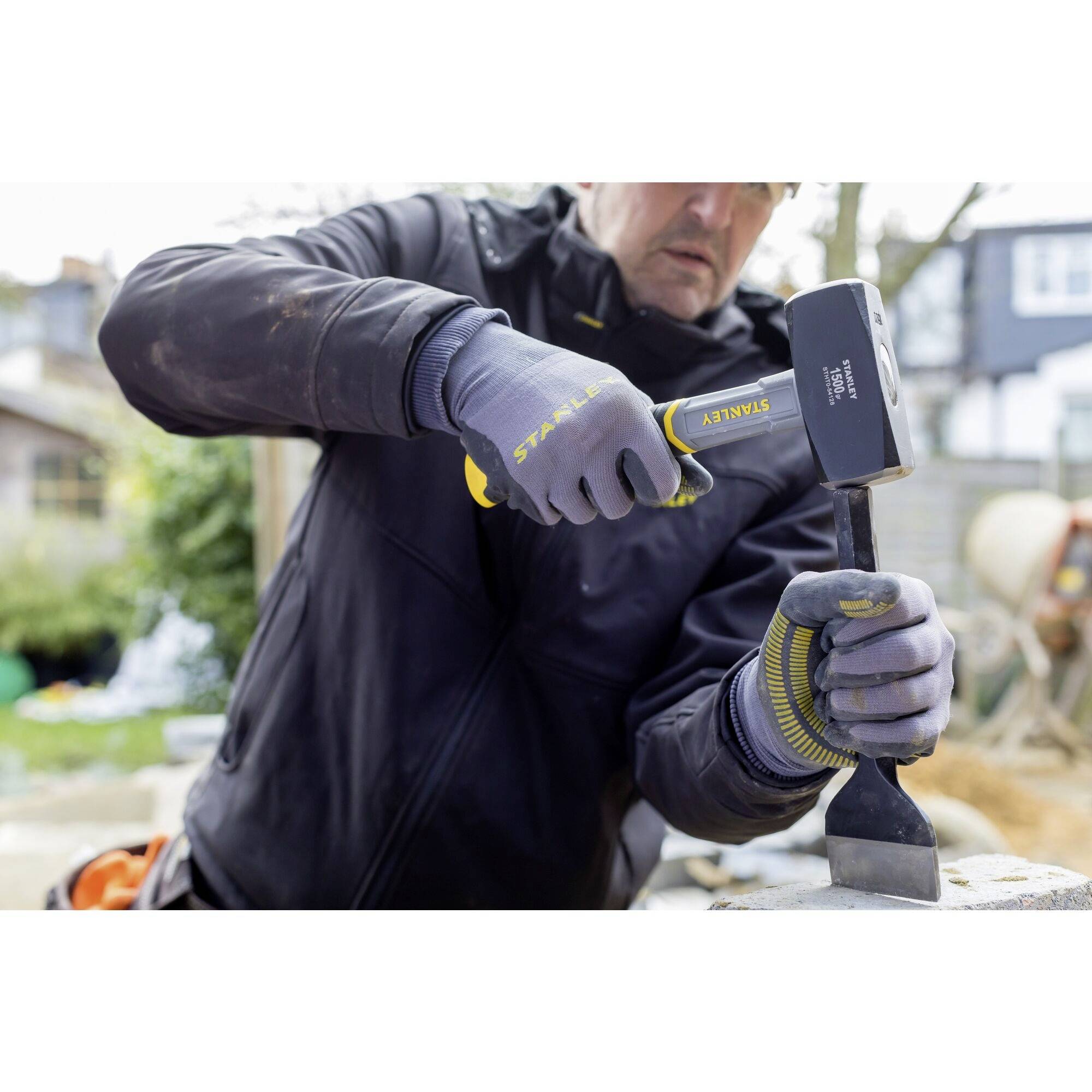 A person is working with a hammer and chisel on a stone. The person is wearing work gloves and a jacket. It is a construction environment.