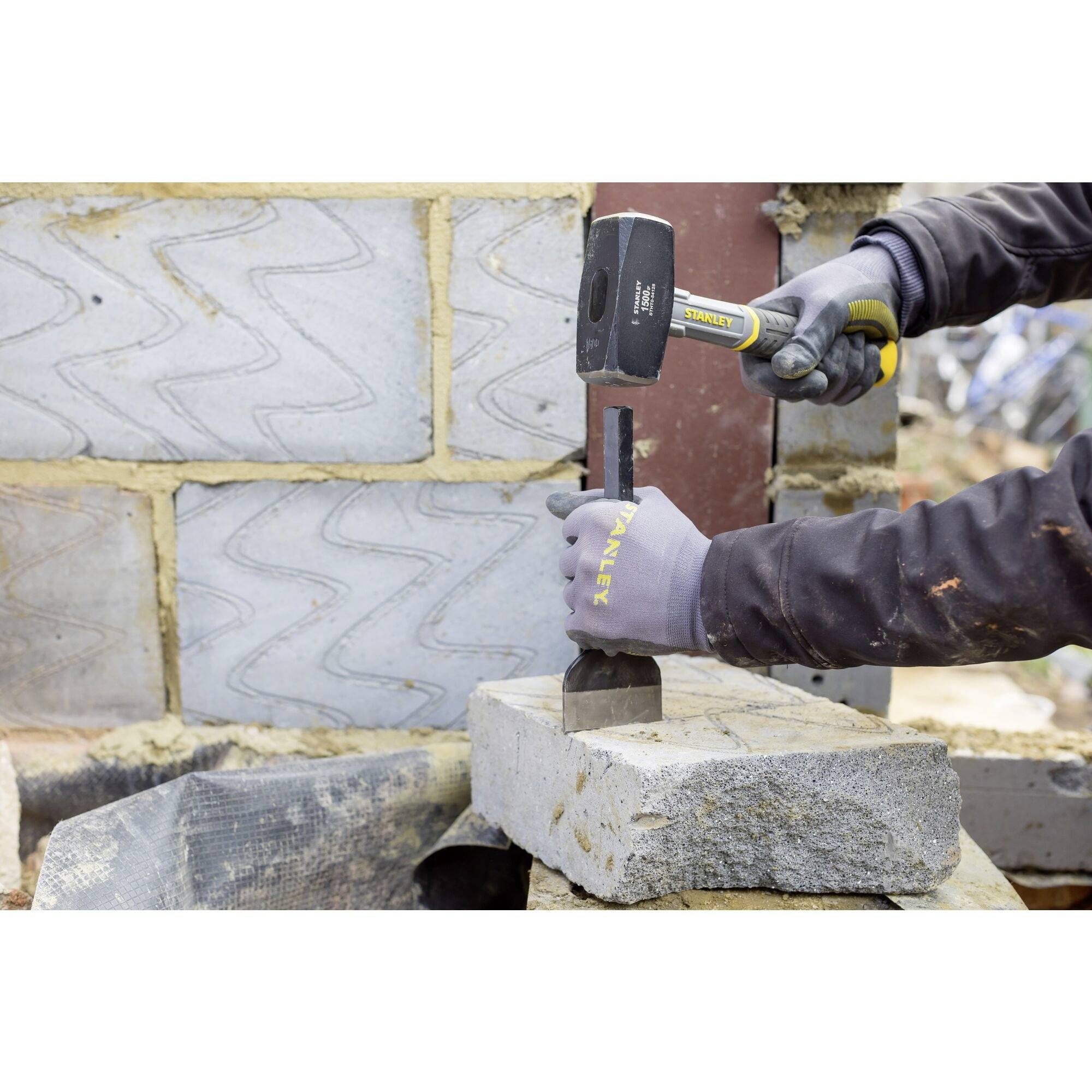A person is chiselling a stone slab with a hammer and chisel. The wall in the background is made of unfinished bricks.