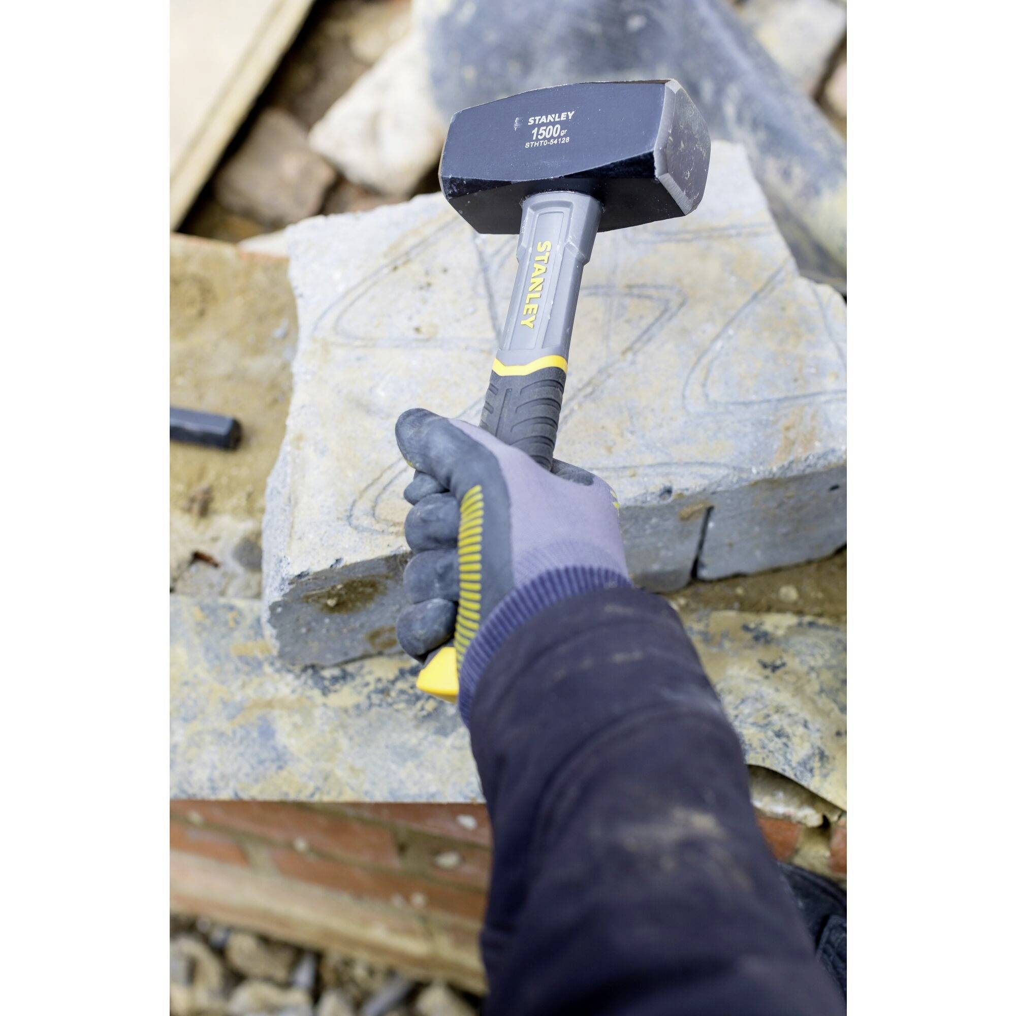 A person wearing a glove holds a heavy hammer over a rock block with an etched pattern, poised to strike.