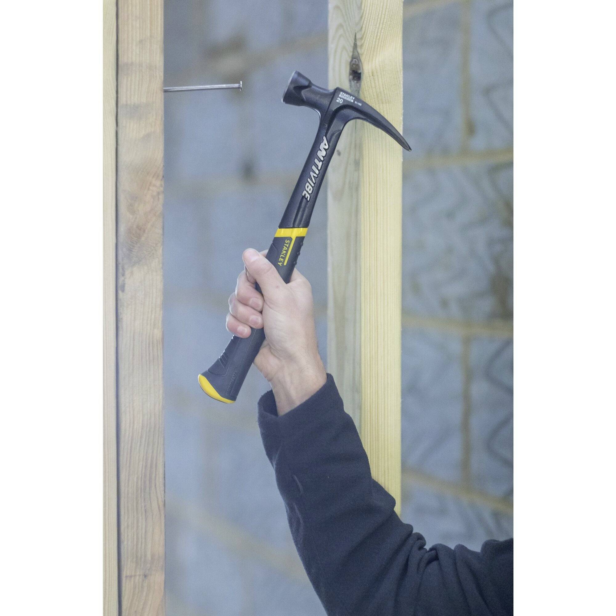 A person is hammering a nail into a wooden board. A brick wall is visible in the background.