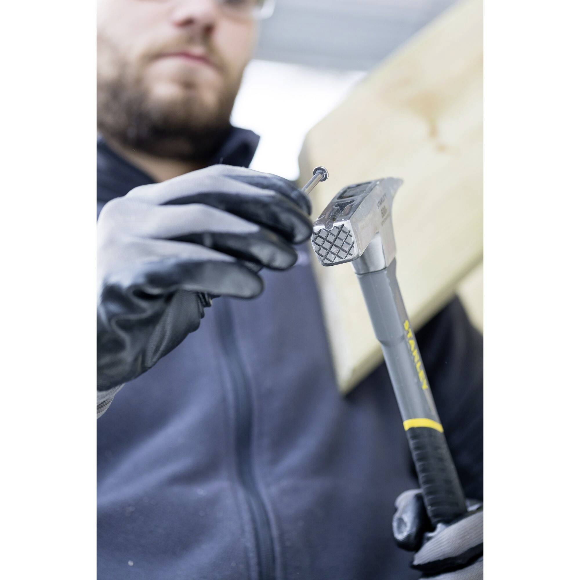 A person in work attire is holding a hammer and a nail, ready to strike into a piece of wood.
