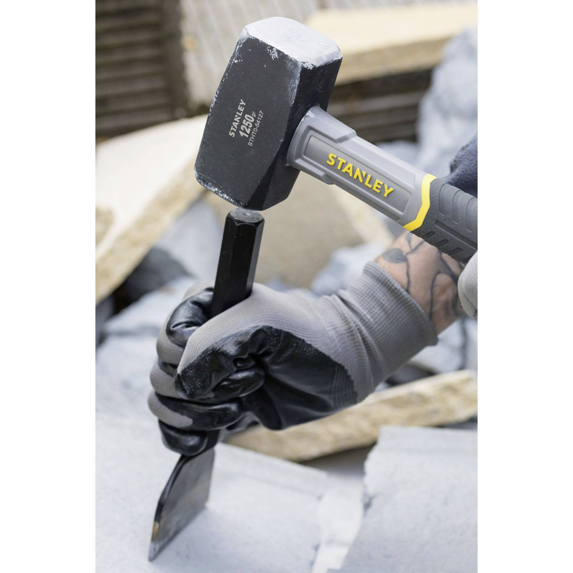 A person is using a black hammer and chisel to work on a grey stone. Wearing work gloves.