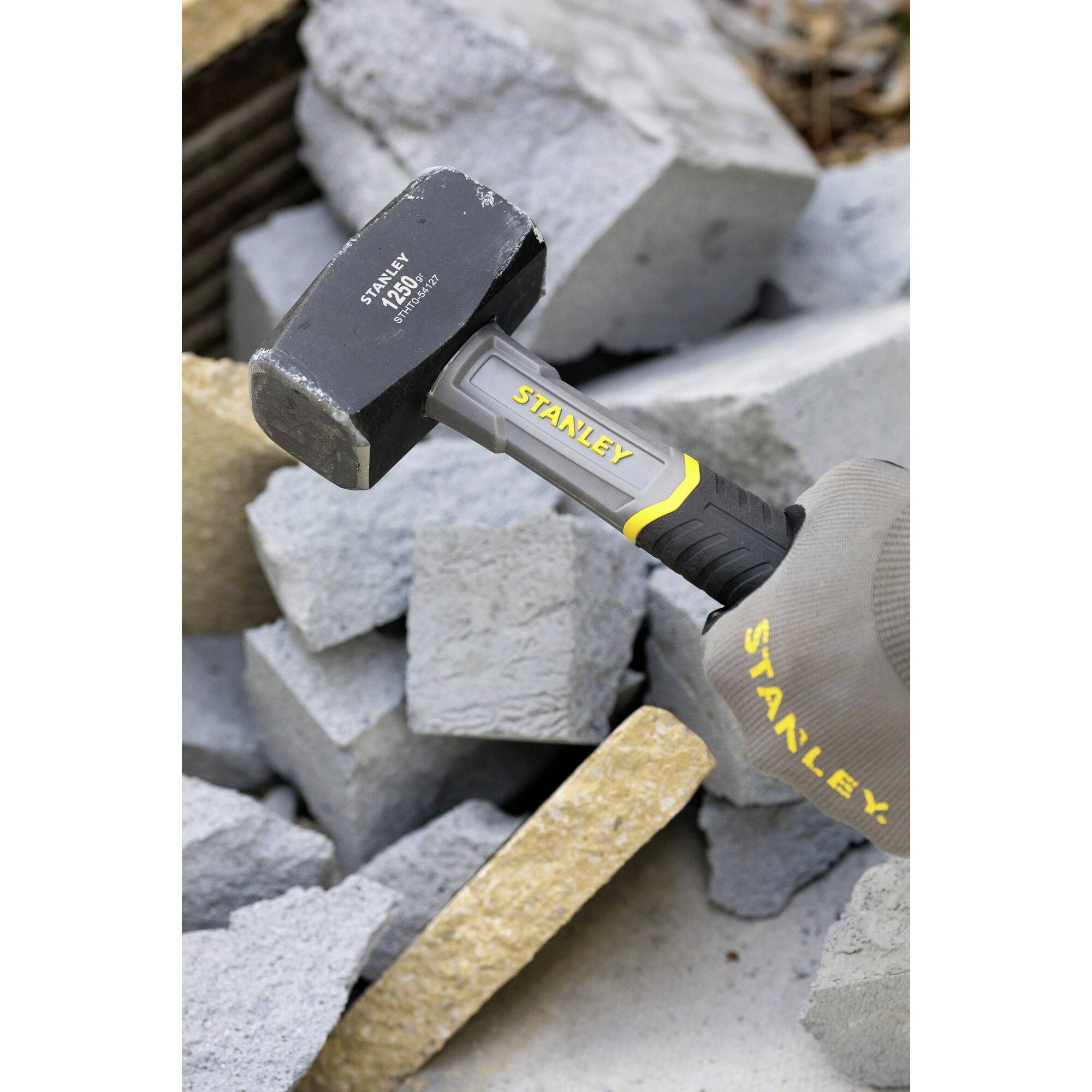 A hand holds a Stanley hammer over a stack of broken concrete blocks. The hammer stands out through its yellow lettering.