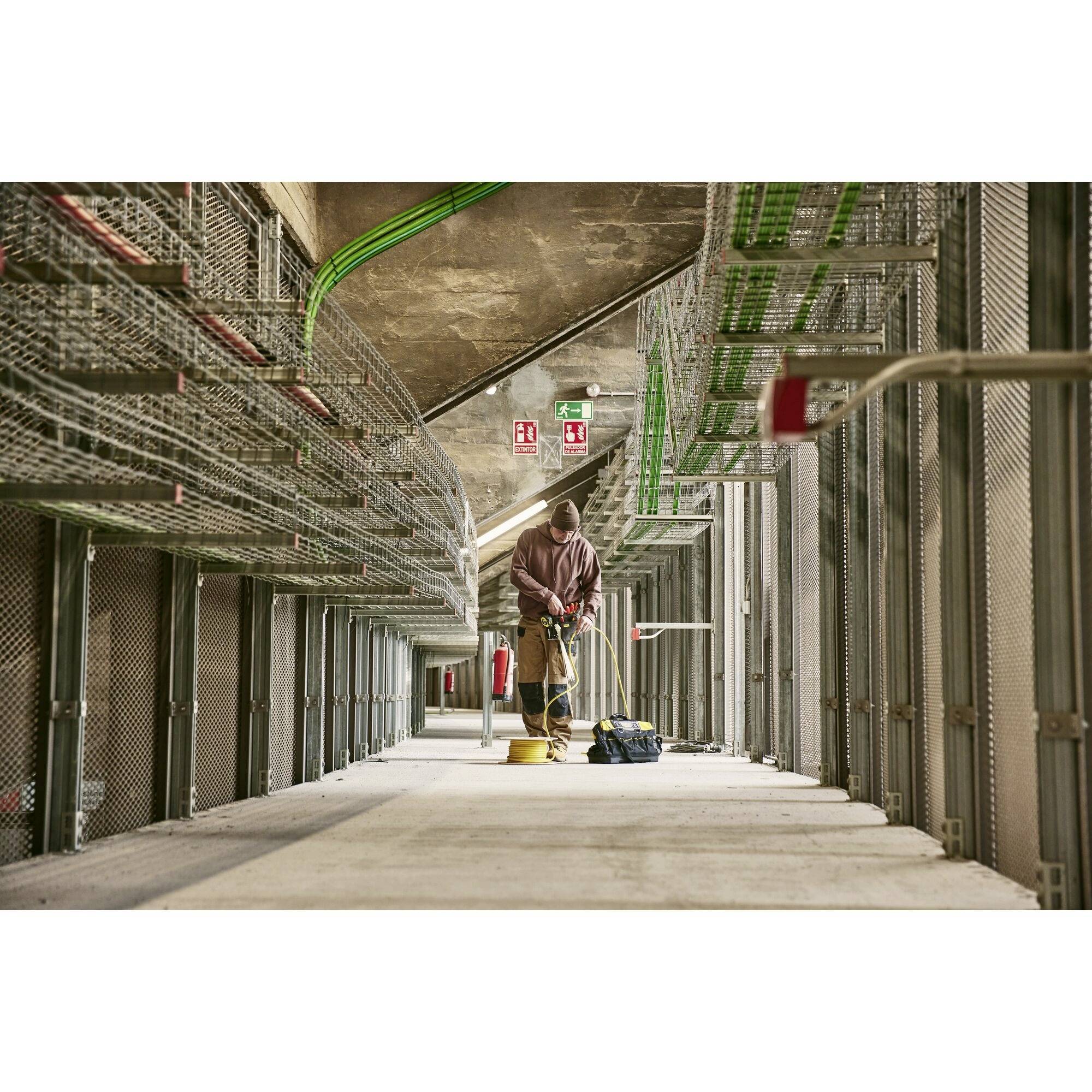'A worker in protective clothing is cleaning a construction site area. Cable trays and pipework are visible overhead.'