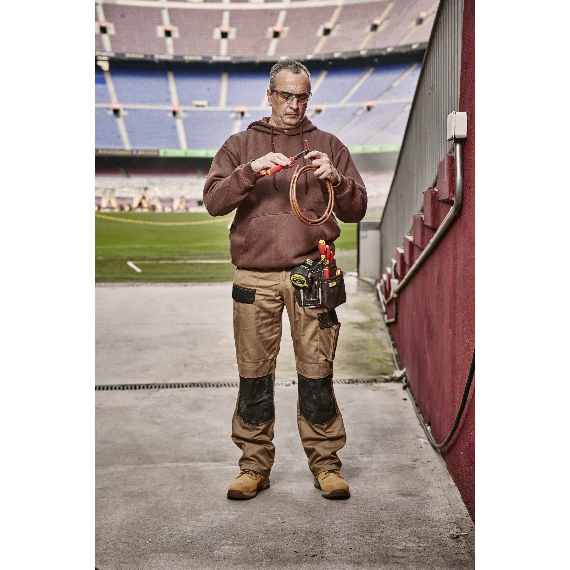 A tradesman stands in a stadium, holding cables in his hands, ready to work. He is wearing work attire and carrying tools.