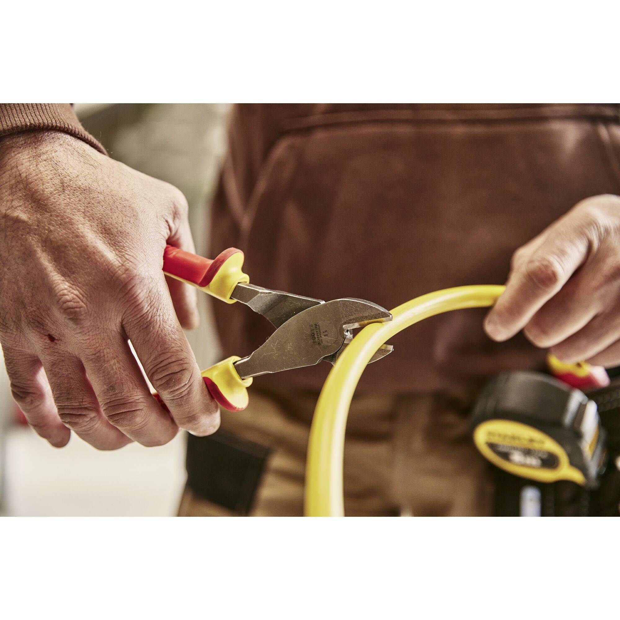 Two hands are cutting a yellow cable with pliers. A tape measure on a belt clip is visible in the background.