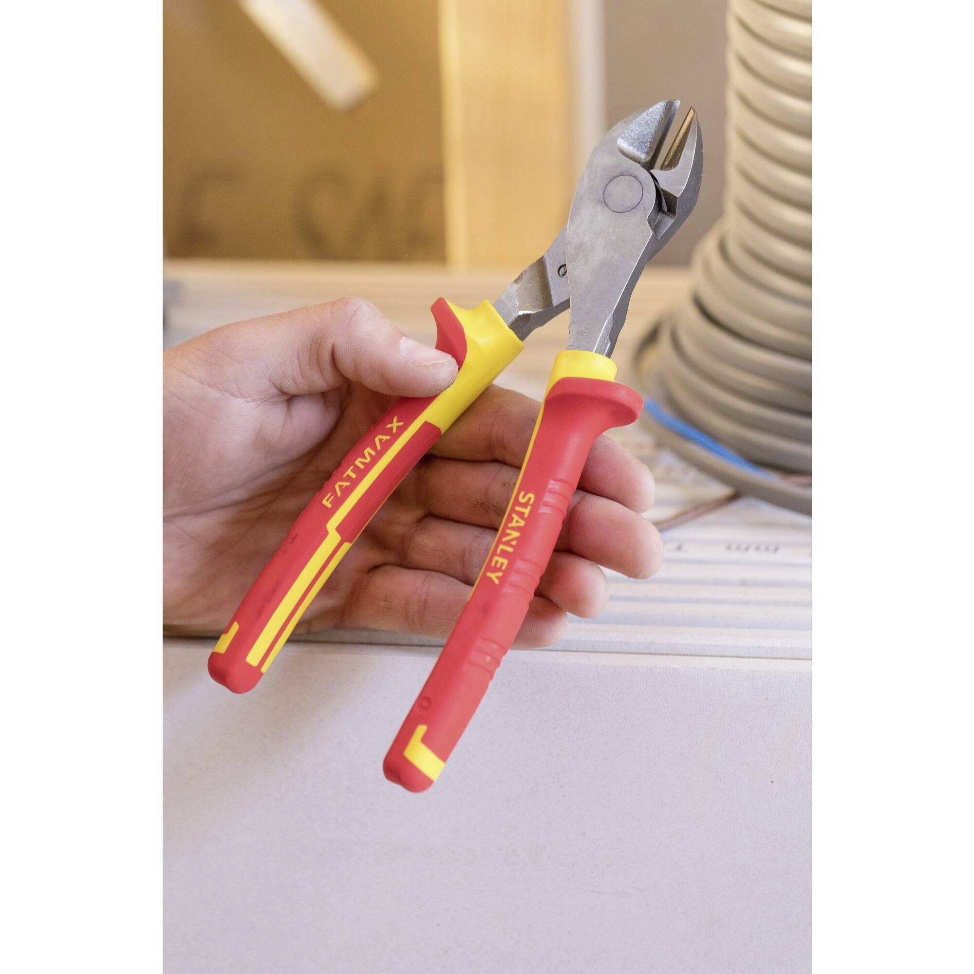 A hand is holding wire cutters with red and yellow handles in front of a blurred construction site background.