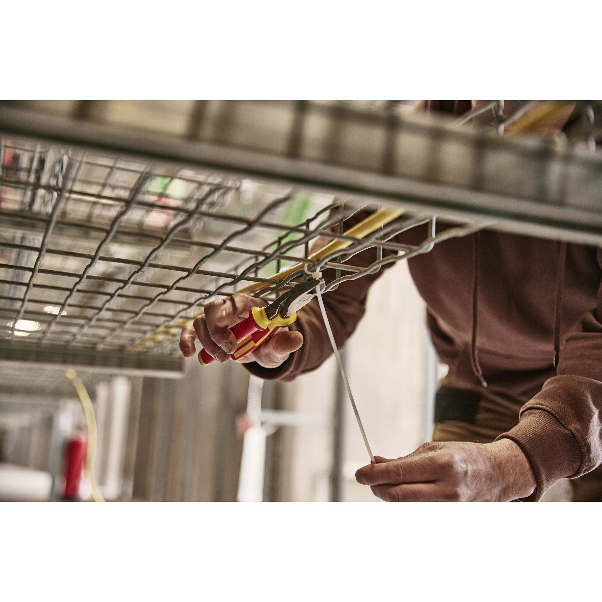 A person is installing electrical cables on a metal grid. The person is wearing brown clothing and holding tools in their hands.