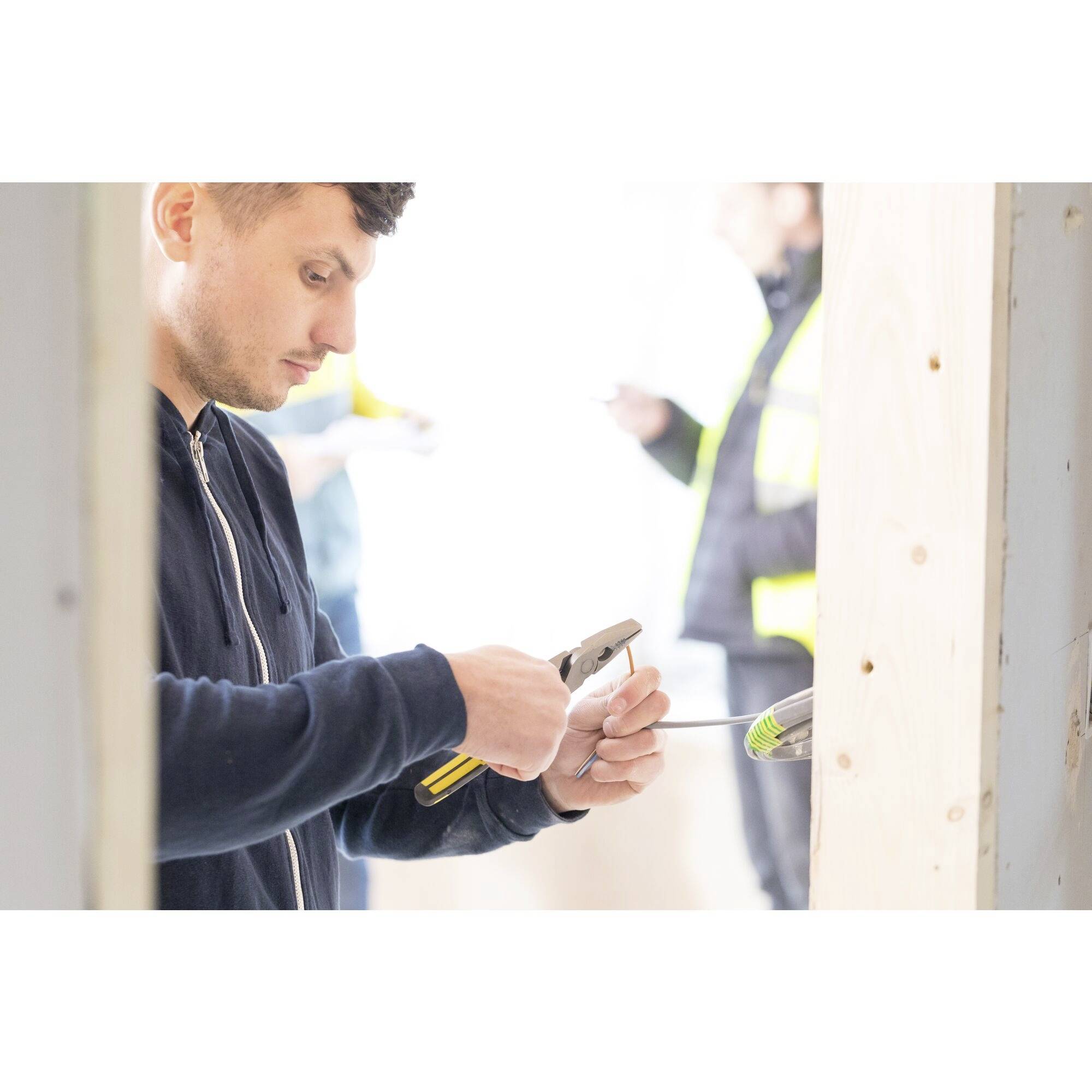 A man in workwear is holding a hammer and hammering a nail into a wooden frame, with a blurred worker visible in the background.
