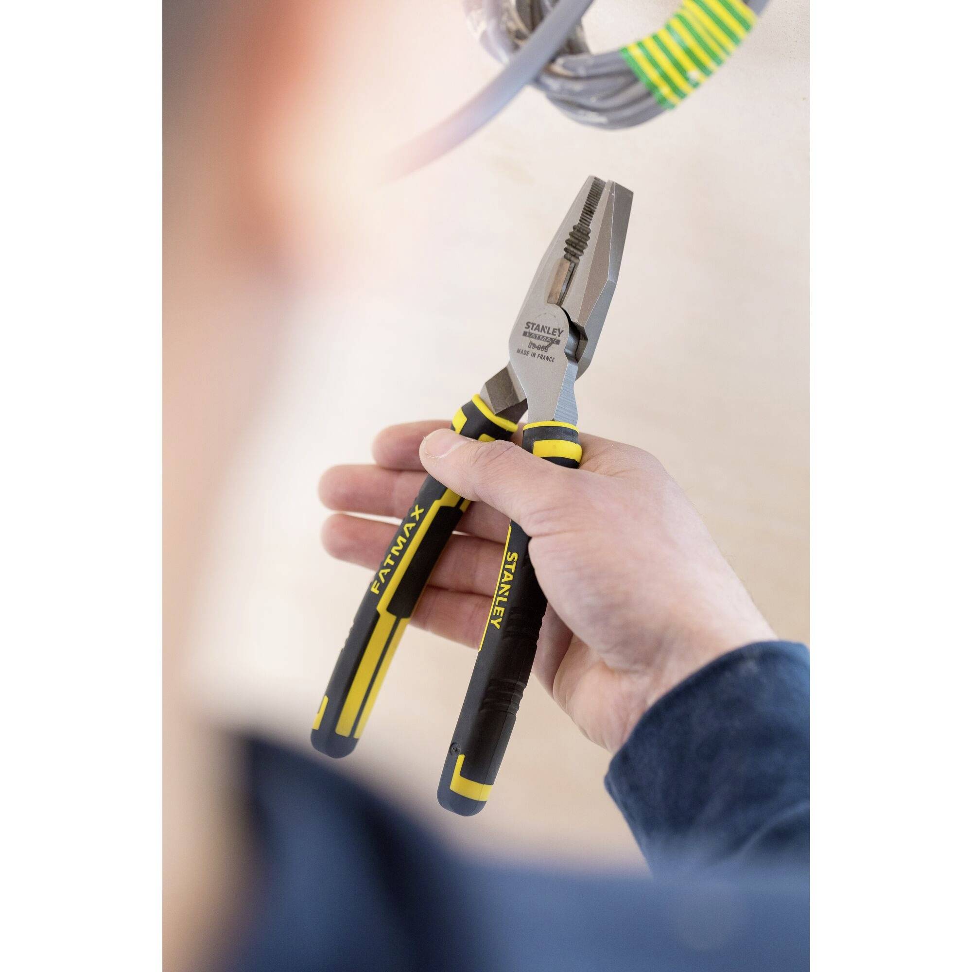 A hand is holding a yellow and black combination pliers to work on a cable. A grey cable is visible in the background.