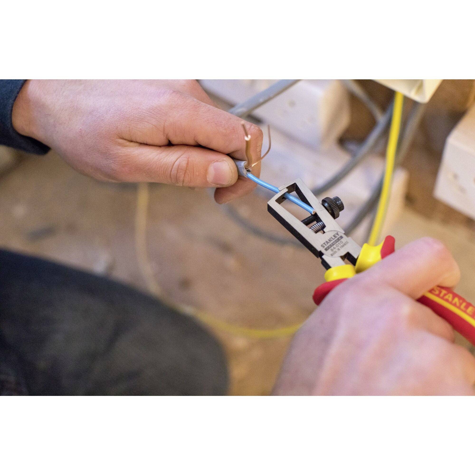 A person is using a wire stripping tool to expose a cable. Plug sockets can be seen in the background.