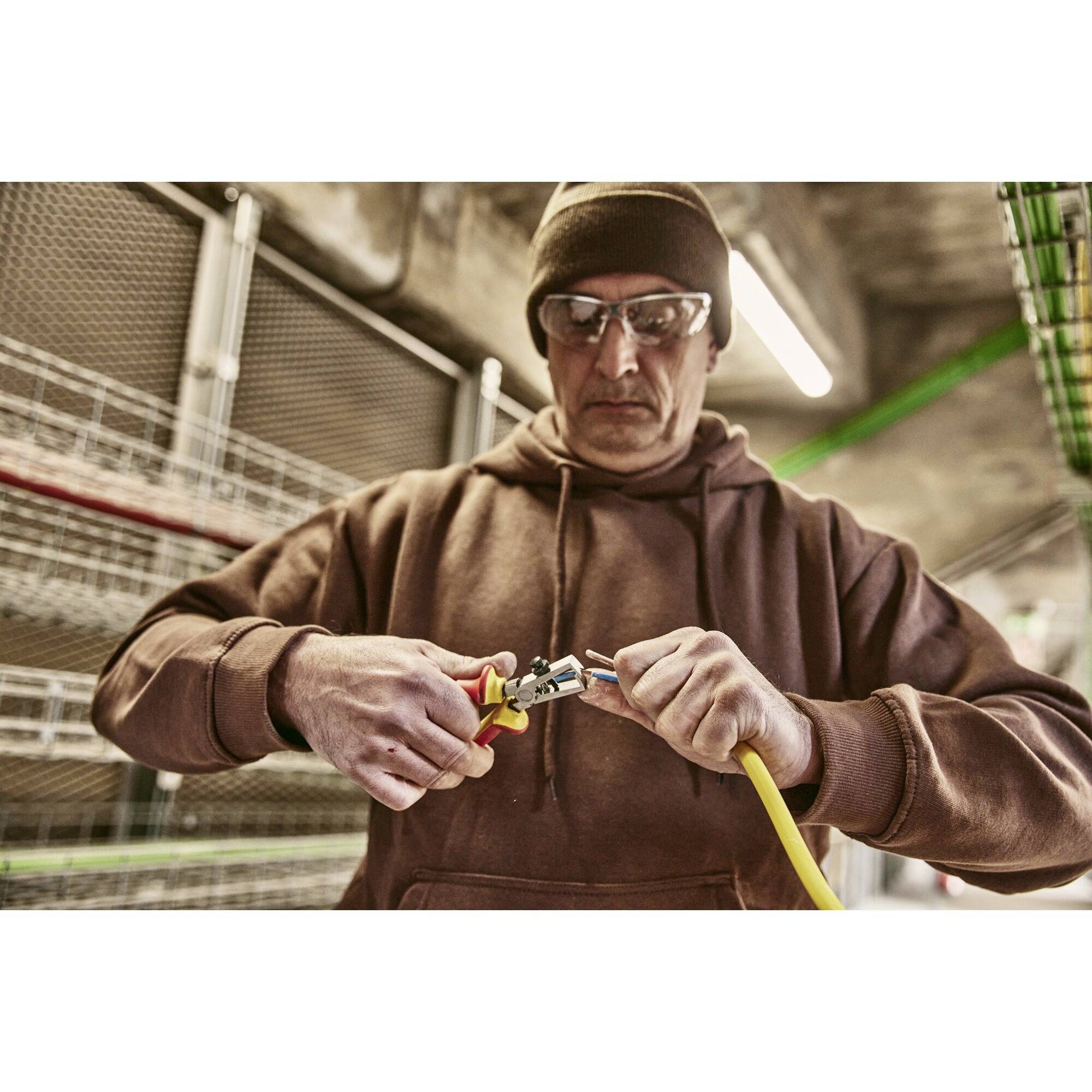 A middle-aged person wearing safety glasses and a cap is working in an industrial environment, using wire cutters on a yellow cable.