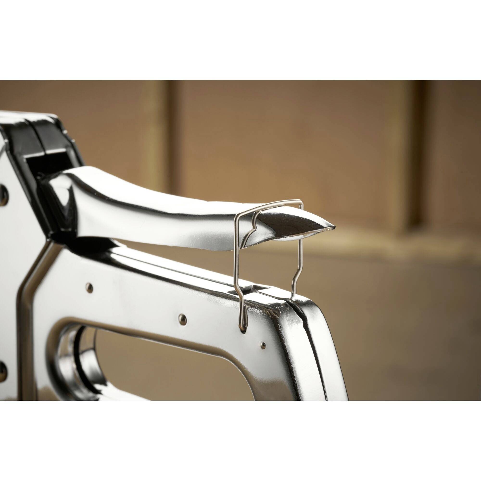 Close-up of a chrome-plated staple gun, positioned on a table with a blurred background. The focus is on the mechanics of the stapler.