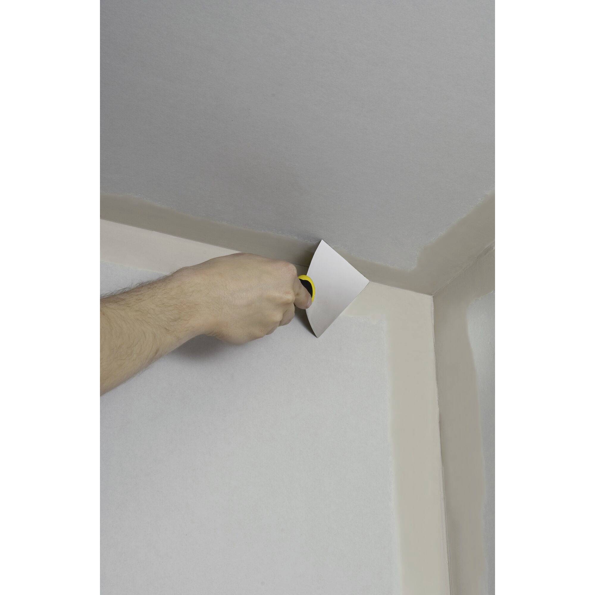 A person is painting white paint at the corner of a wall and ceiling using a paint roller. The wall is partially painted.