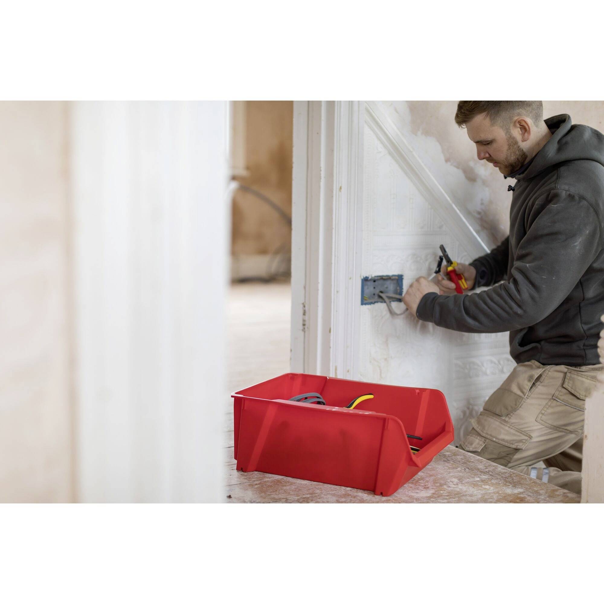 A man is installing a socket on the wall using pliers. A red toolbox sits on the table in the foreground.