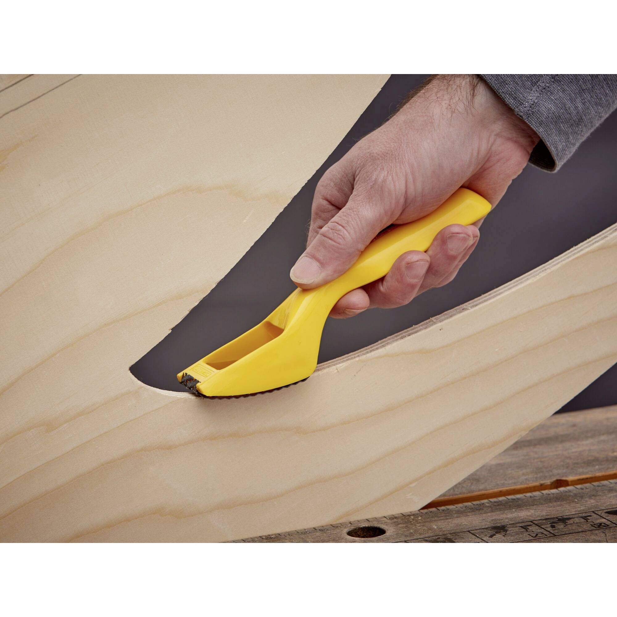 A hand is holding a yellow tool that is smoothing the edges of a wooden board. The board has a curved shape.