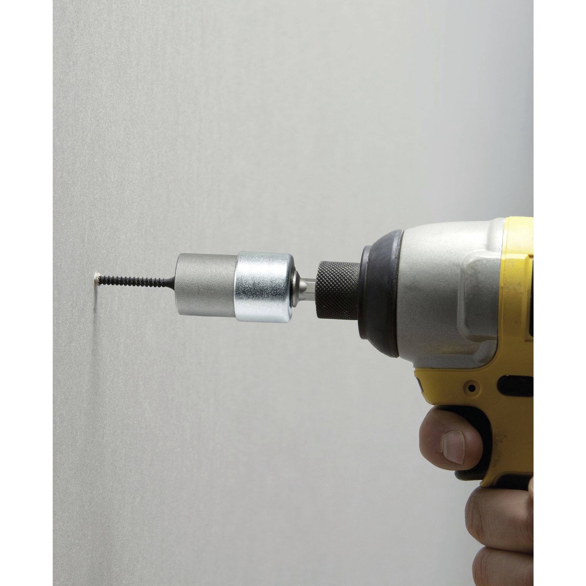 A cordless drill drives a screw into a white wall. A metal adapter surrounds the screw for precise work. A hand supports the tool.