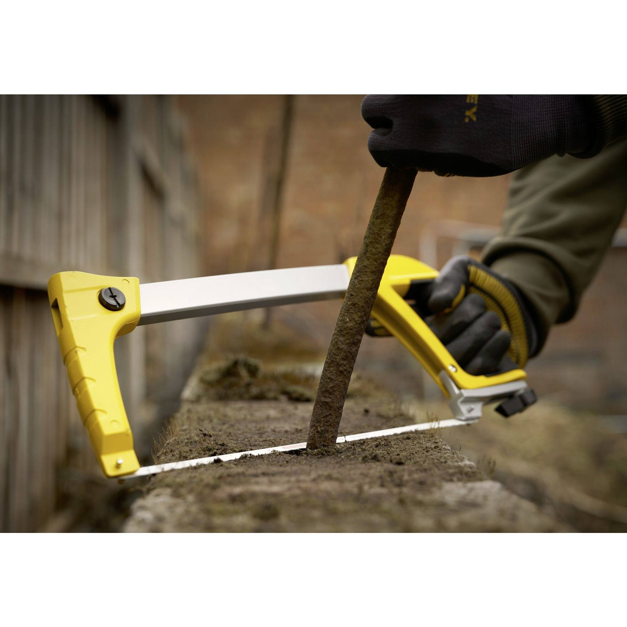 A person is sawing a thin branch with a yellow handsaw. The surroundings appear rural with wooden fences in the background.