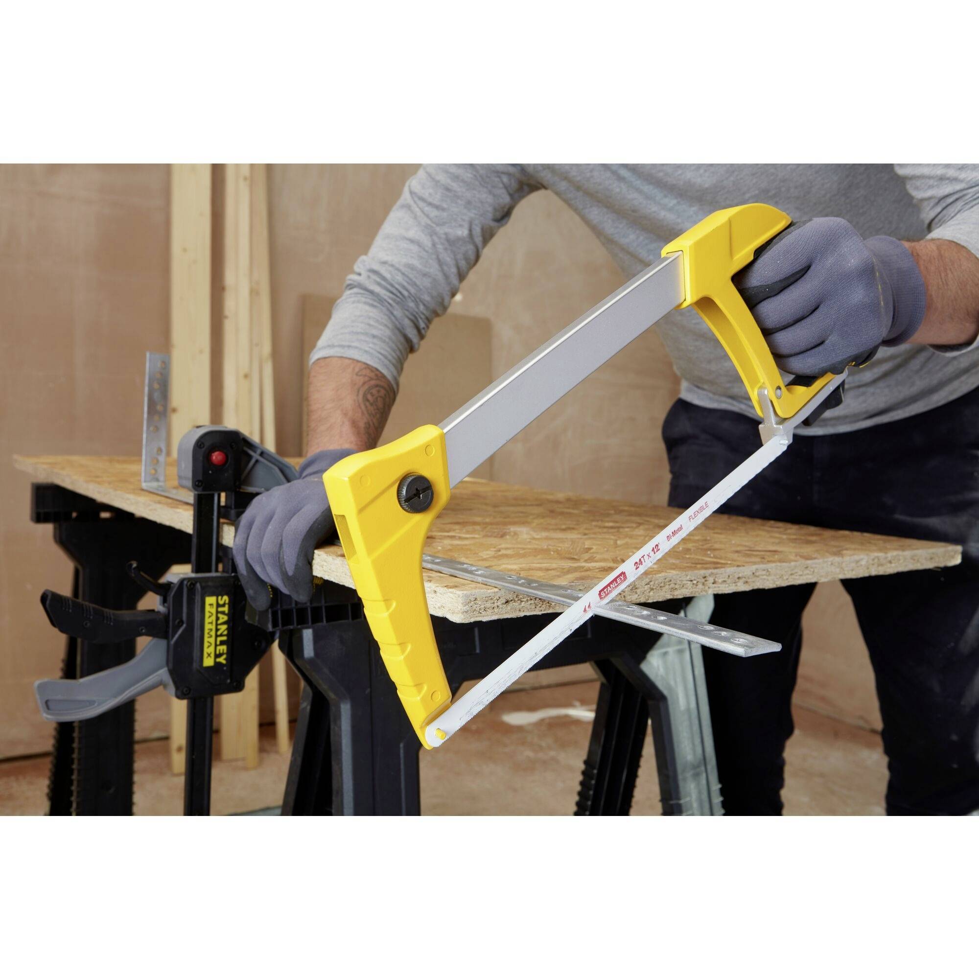 A person is sawing a wooden board, which is resting on two trestles, with a yellow metal saw in a workshop setting.