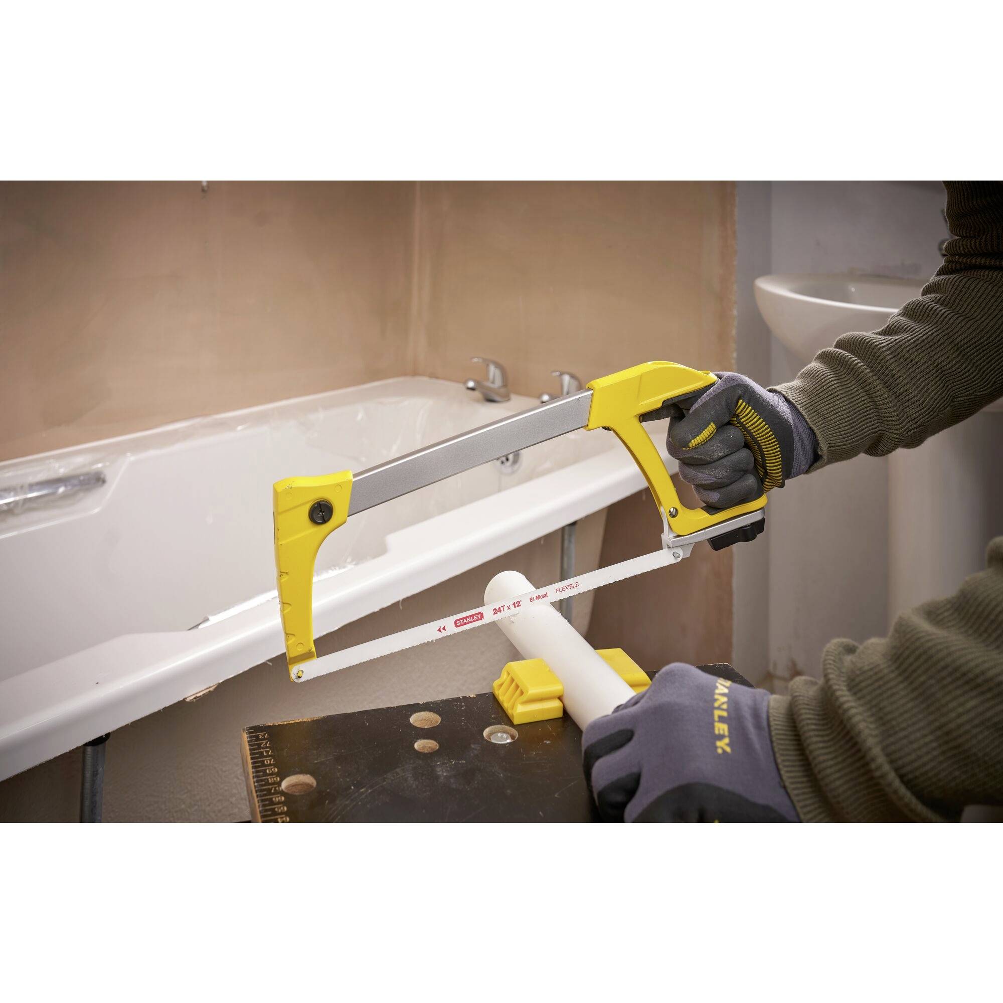 A person is sawing through a pipe in the bathroom using a metal saw. They are wearing gloves. A bathtub is visible in the background.